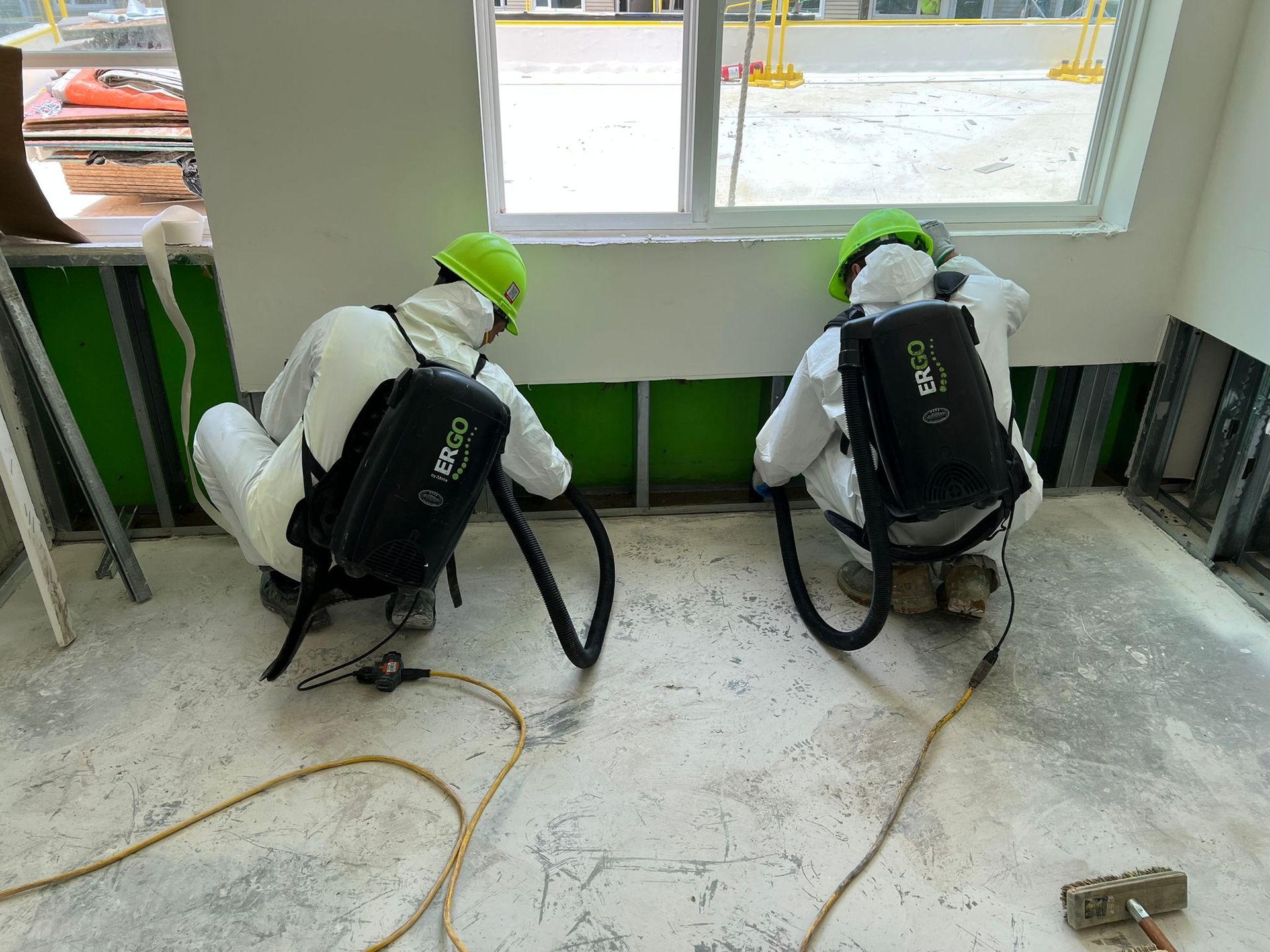 Two workers in protective suits vacuuming a wall, interior setting.