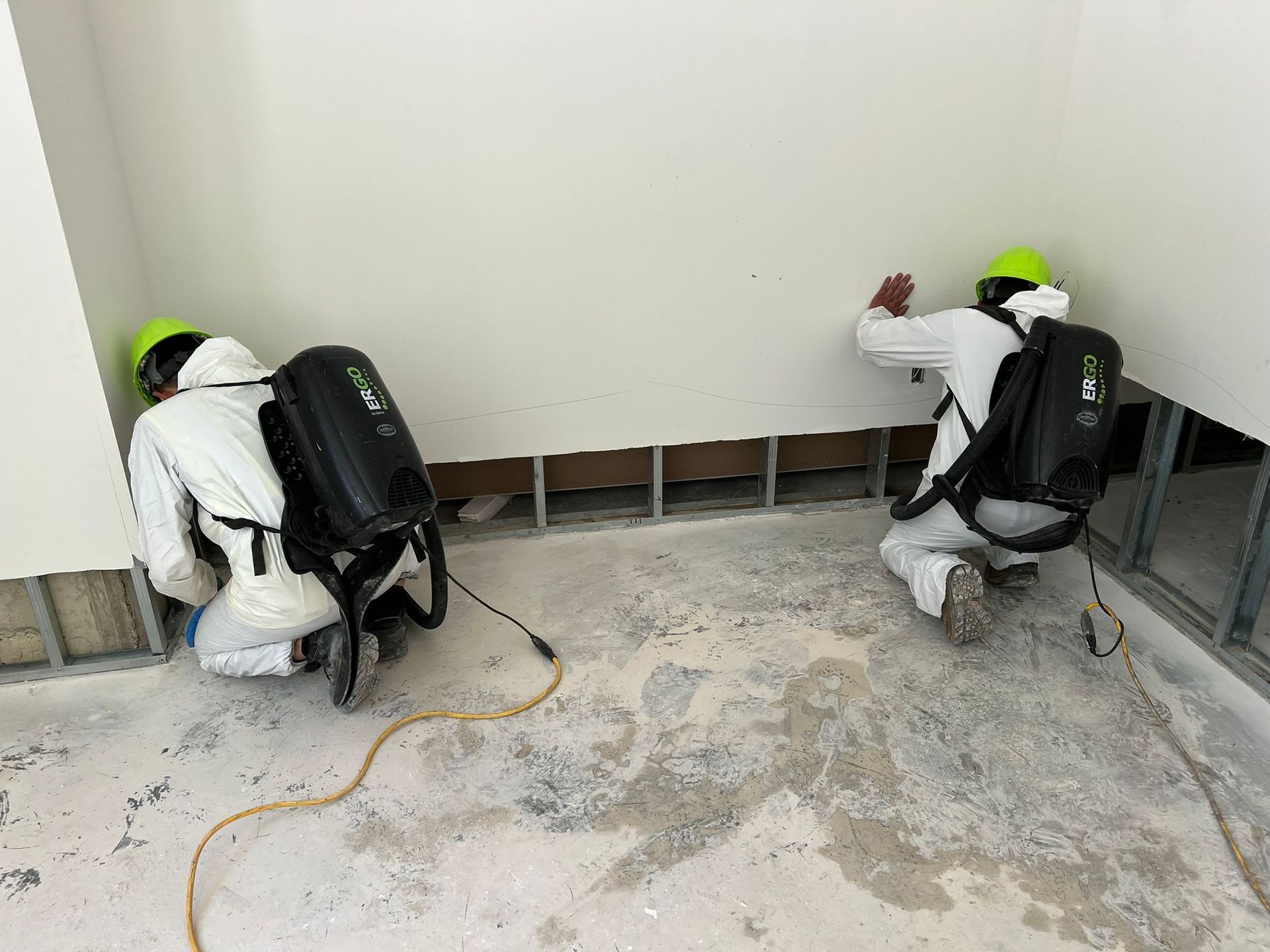 Two workers in protective suits spray a wall. White suits, green helmets, black backpack sprayers. Indoor, unfinished space.