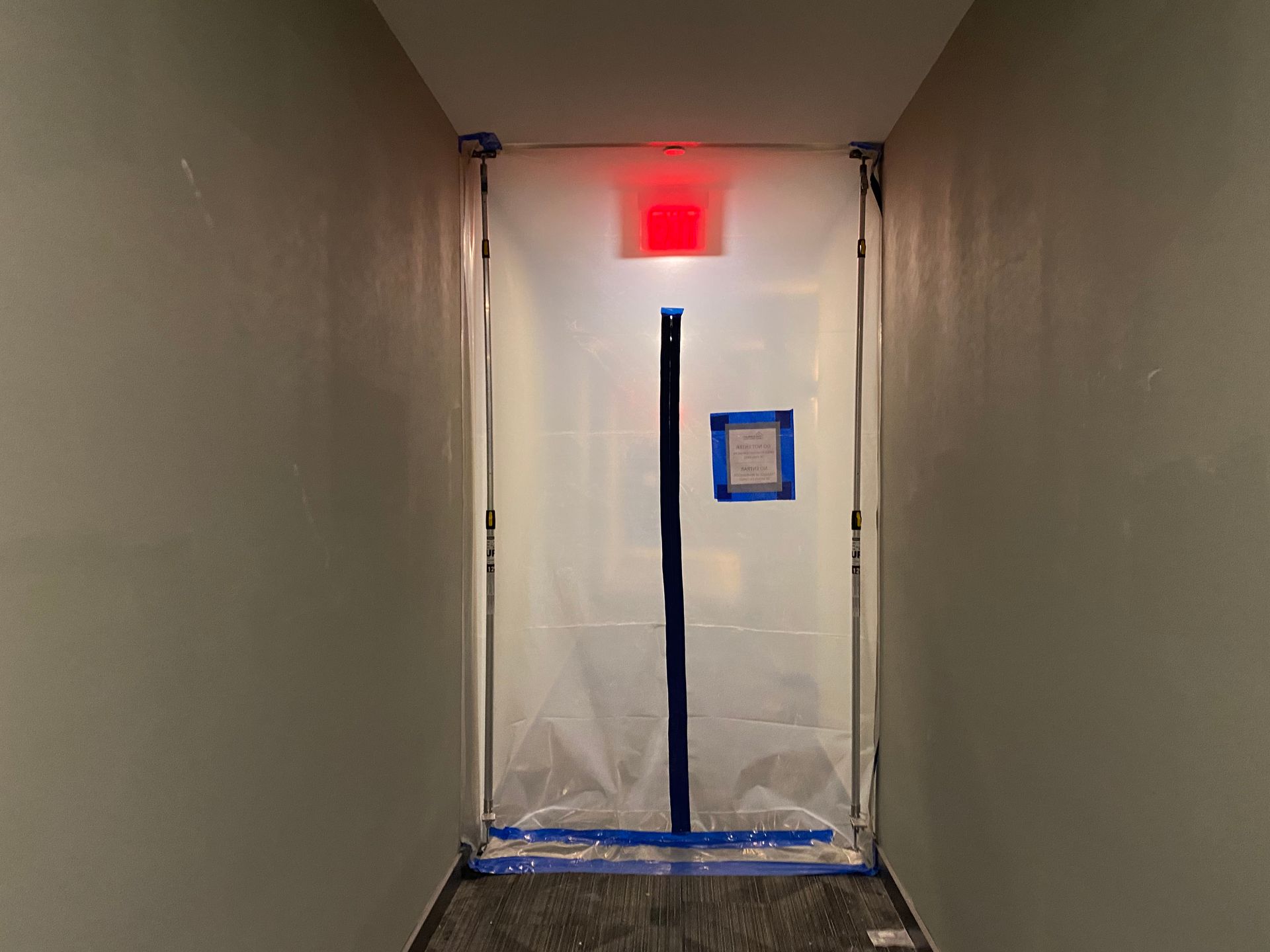 Hallway with doorway sealed off with plastic sheeting; red emergency light above.