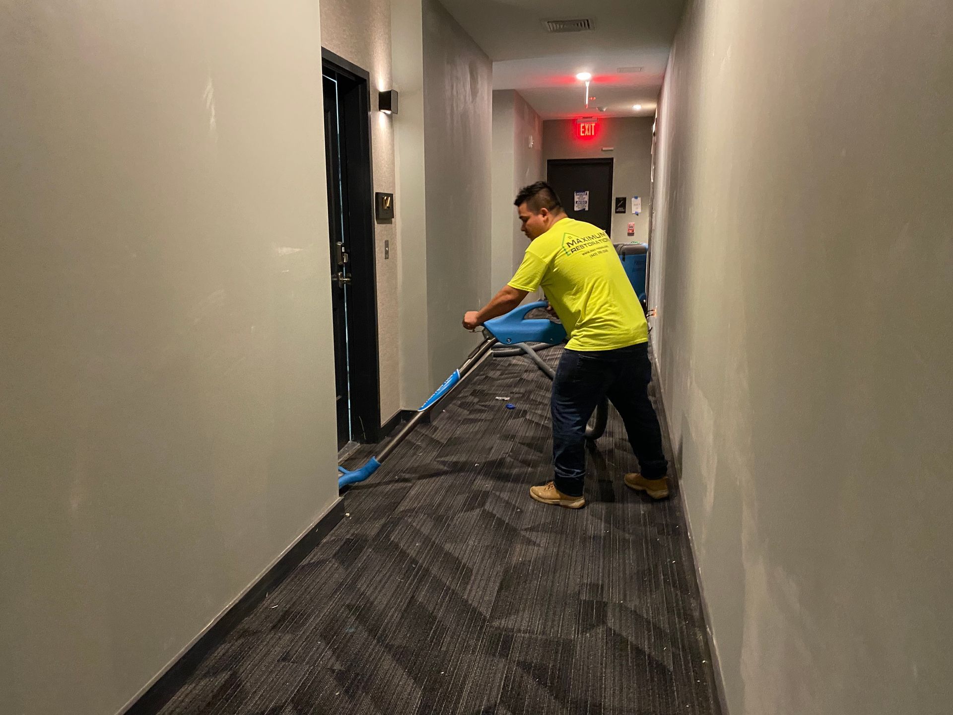 Person vacuuming a carpeted hallway. The worker wears a yellow shirt and dark pants.