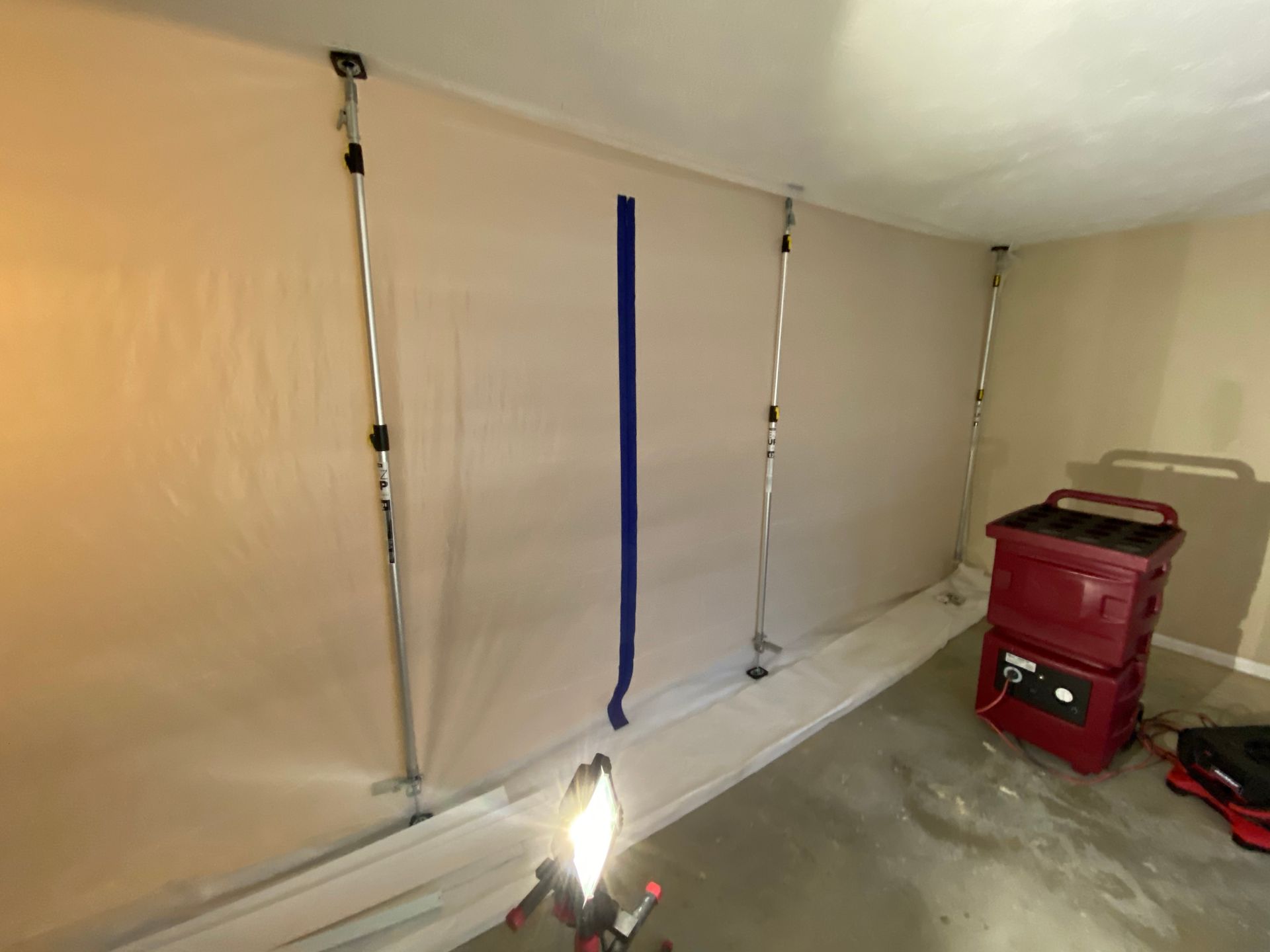 Plastic sheeting covering a wall, held in place by adjustable poles; a work light on the floor.