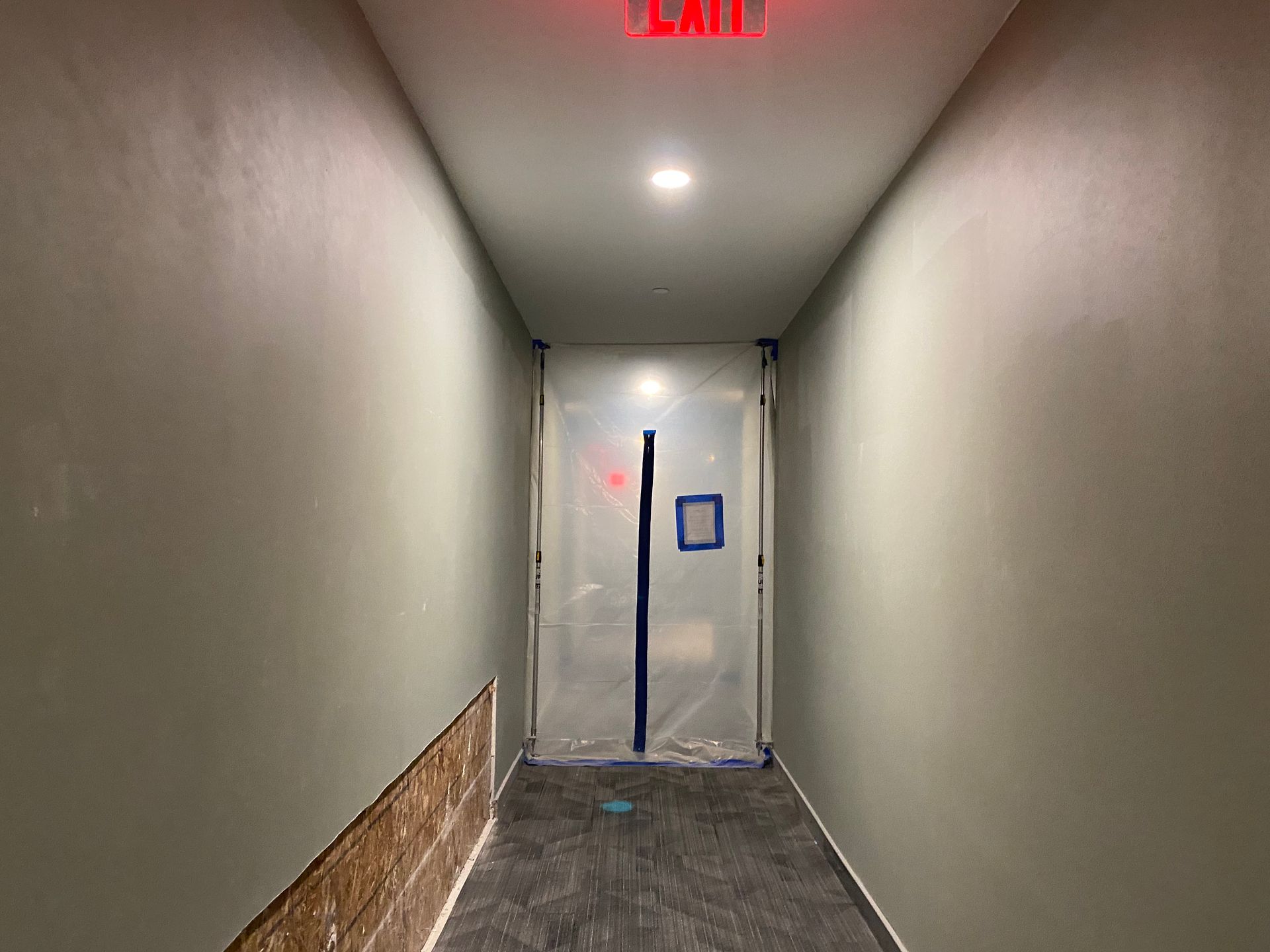 Narrow hallway with exit sign, door covered in plastic, grey walls and floor.