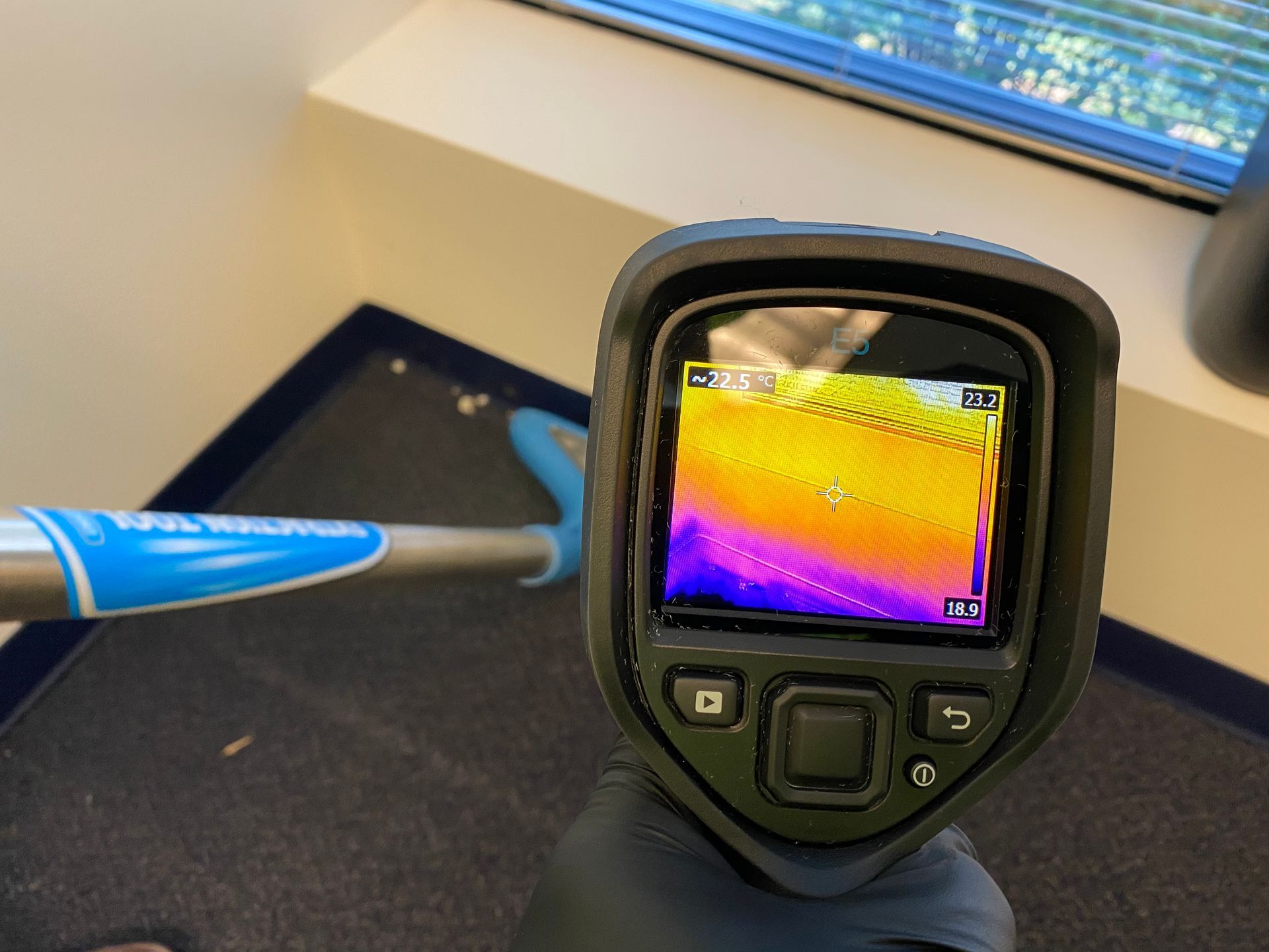 Thermal camera displaying heat readings near a window, held by gloved hand.