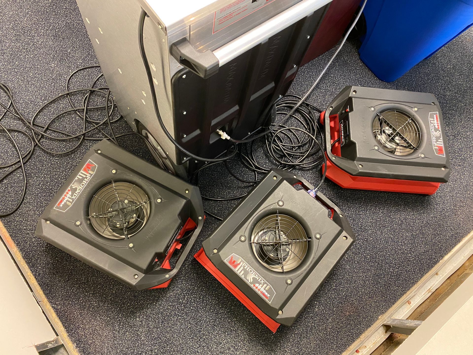 Three red and black air movers on carpet with a large metal box. Cords are tangled.