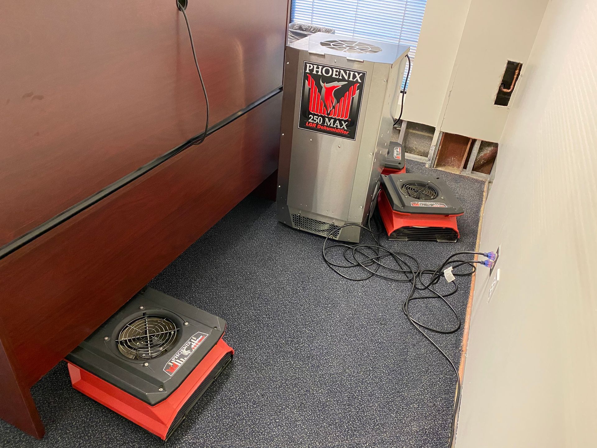 Dehumidification equipment in a room, including a large metal unit and several red and black air movers on a carpeted floor.