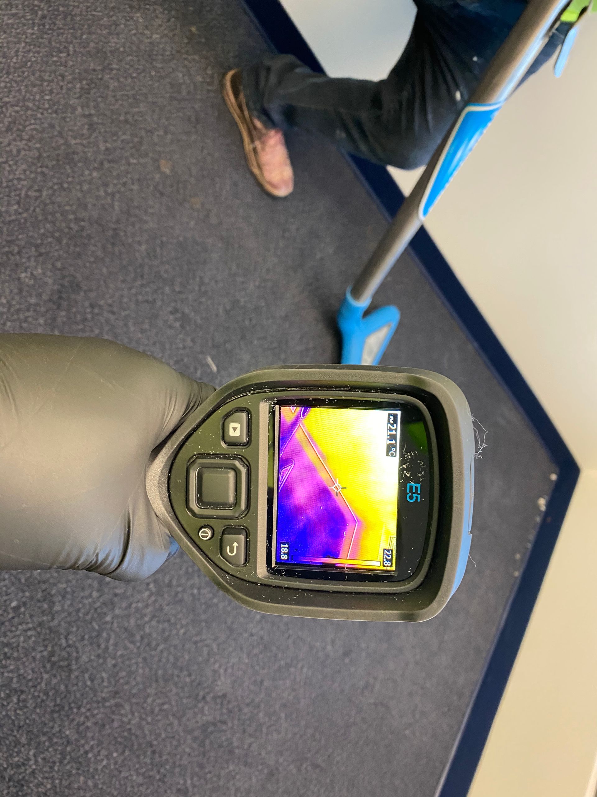 Thermal camera showing color-coded heat map of carpet. Person in background uses a cleaning tool on the carpet.