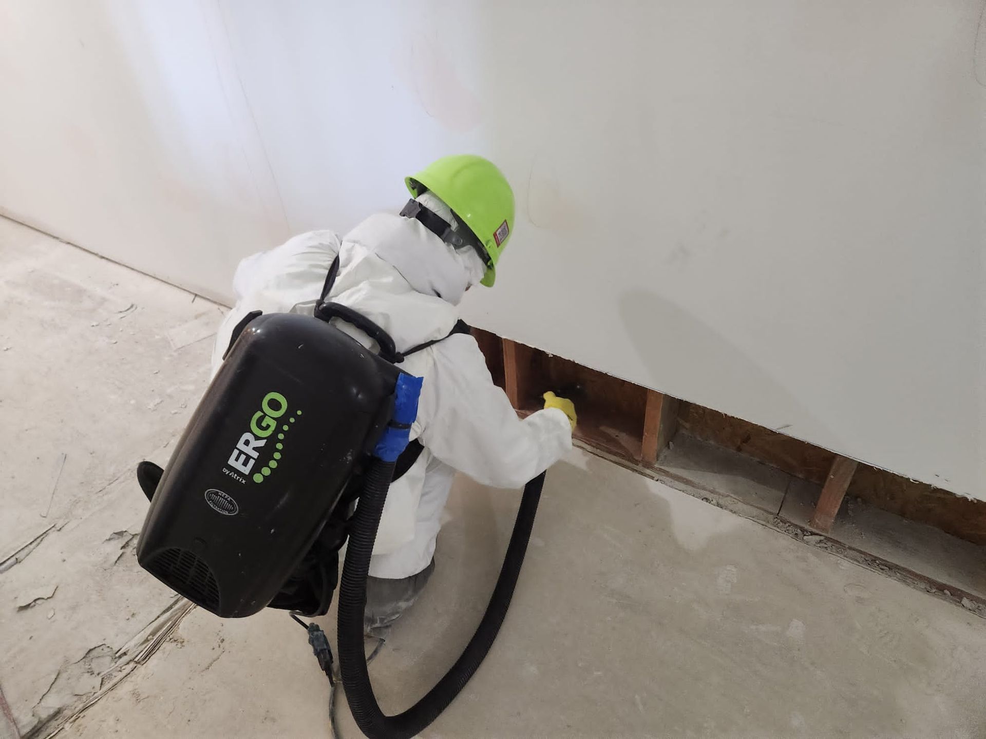 Person in protective gear sprays a wall, with a backpack vacuum, in a construction setting.