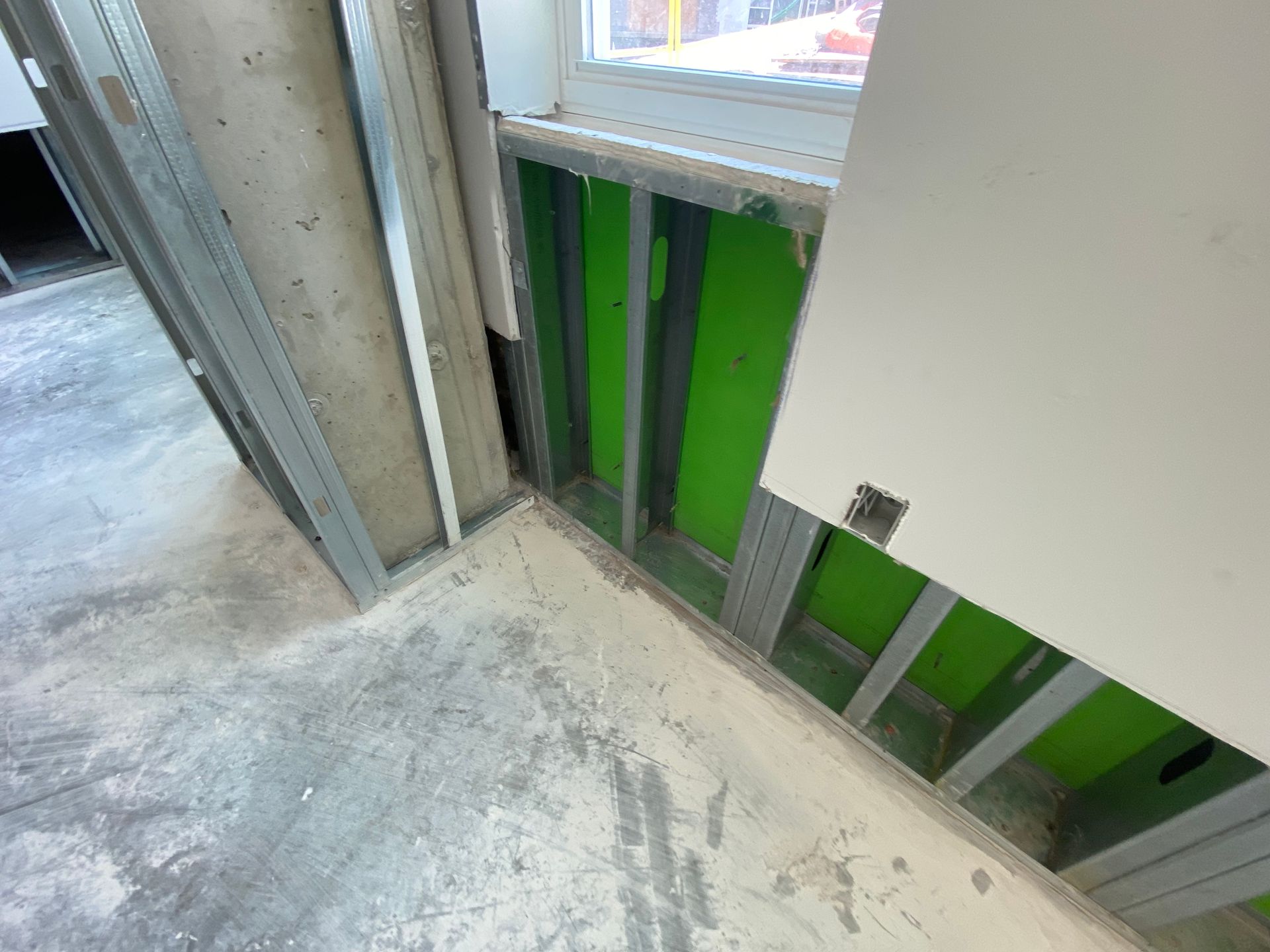Construction site corner with green insulation between metal studs, unfinished walls and floor.