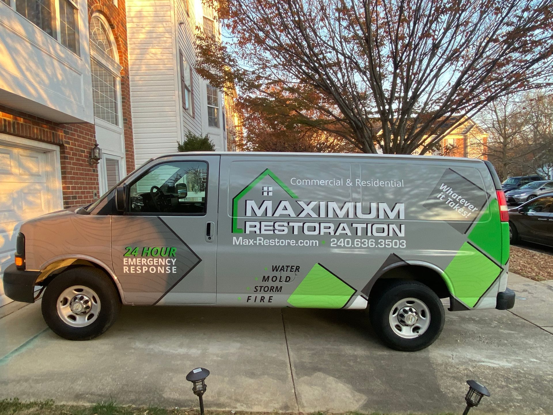 A gray Maximum Restoration work van parked in a driveway featuring green geometric branding, a house logo, and text.