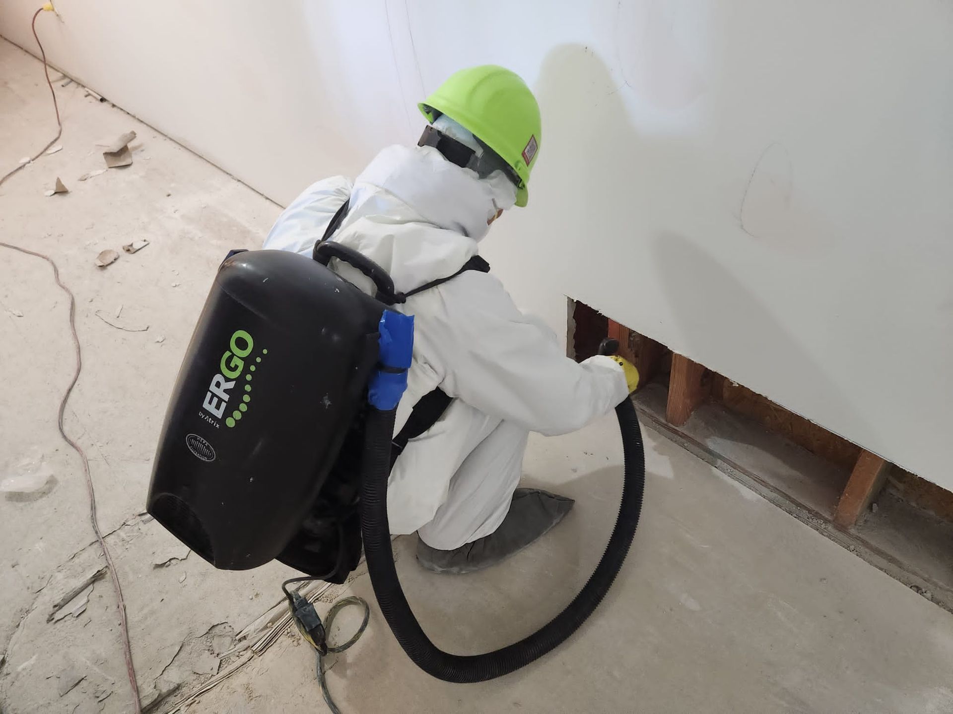 Worker in protective suit using a backpack vacuum on a wall.