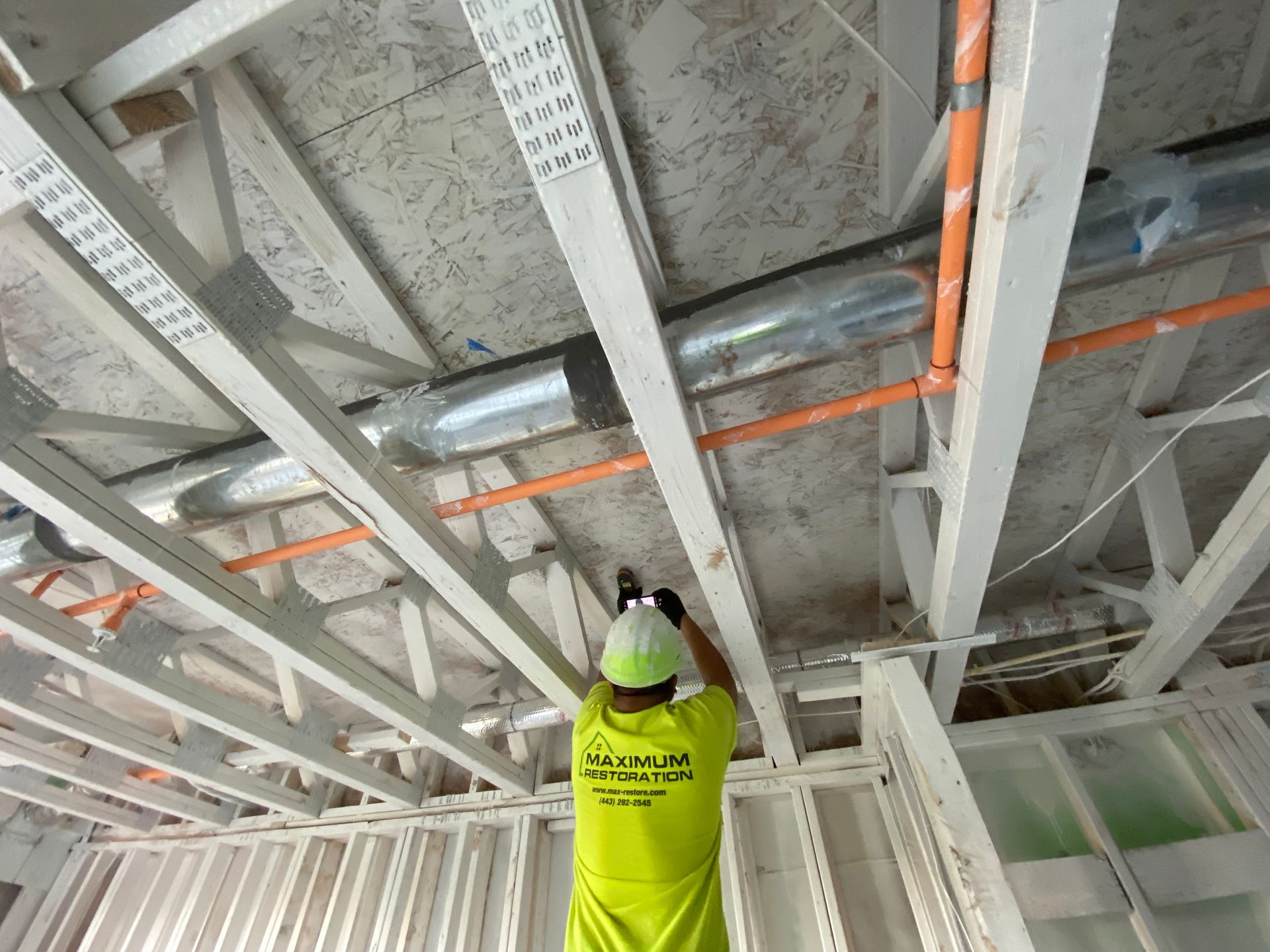 Construction worker installing pipes on a wooden ceiling framework. Worker wears a yellow shirt and a white helmet.