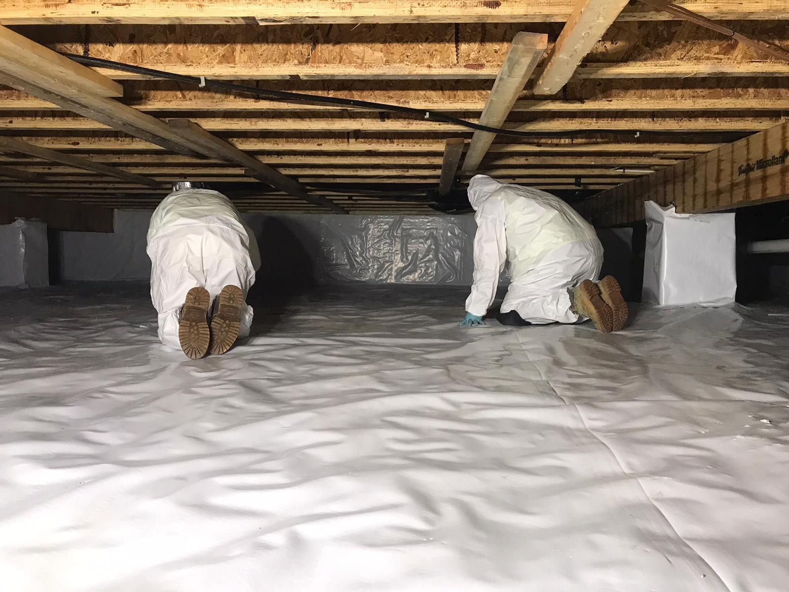 Two people in protective suits crawling in a crawl space, white vapor barrier on floor.