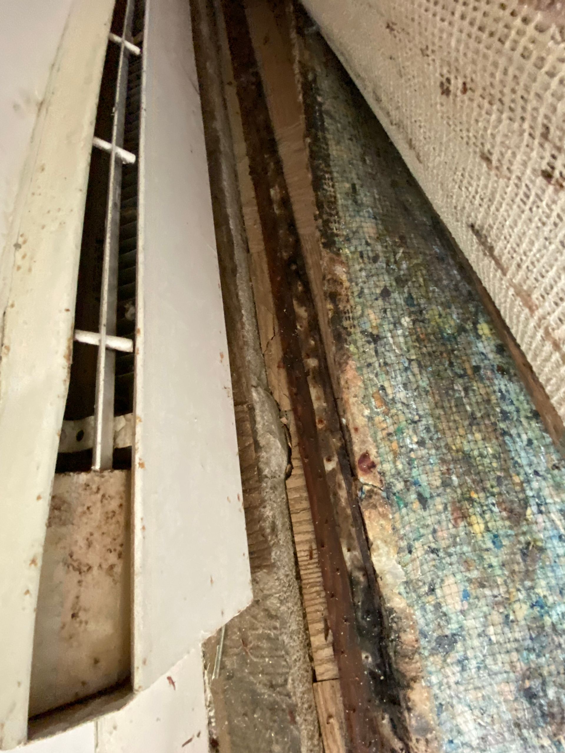 A view of an air vent, a strip of wood and flooring with colorful padding.