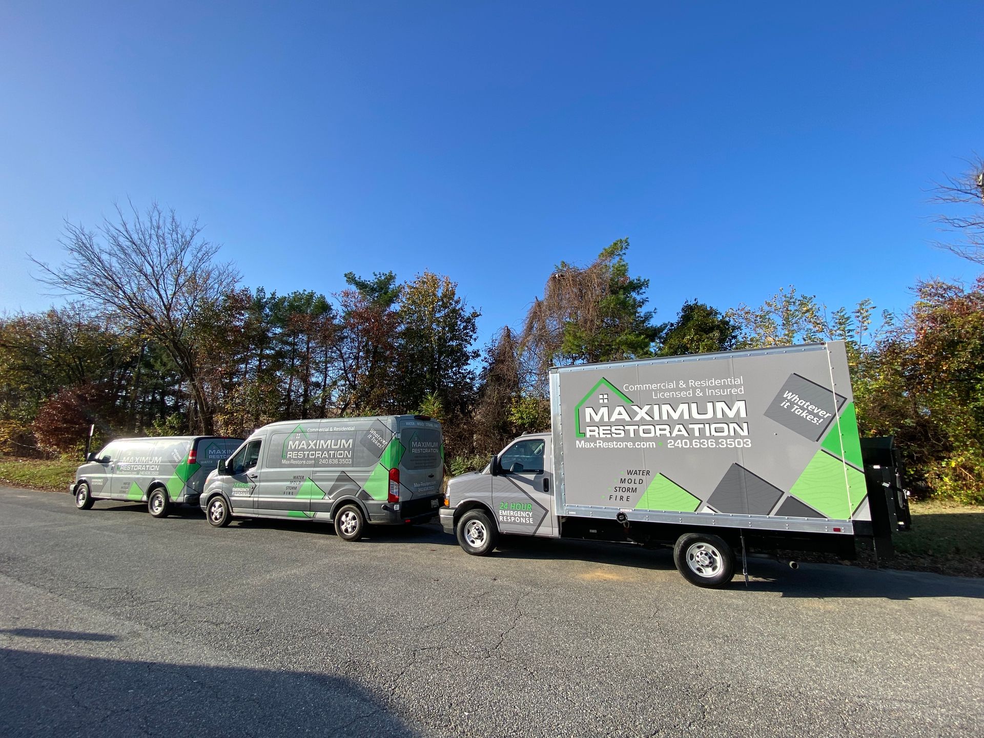 Three Maximum Restoration vehicles parked on a gravel lot under a blue sky, trees in the background.
