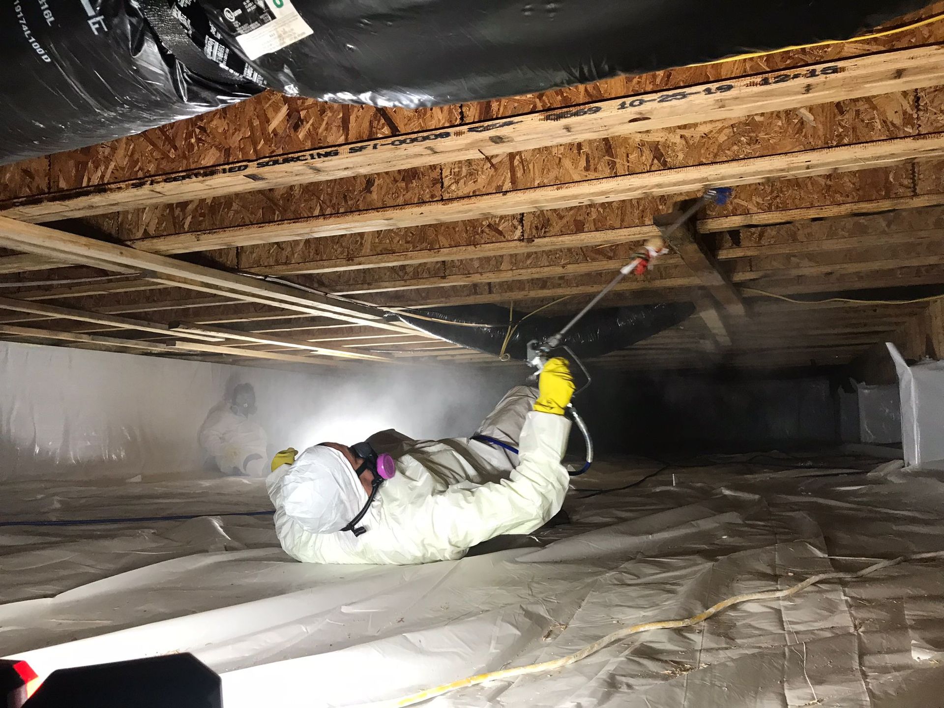 Person in protective suit spraying under a house crawlspace, wooden beams overhead, plastic sheeting on the ground.