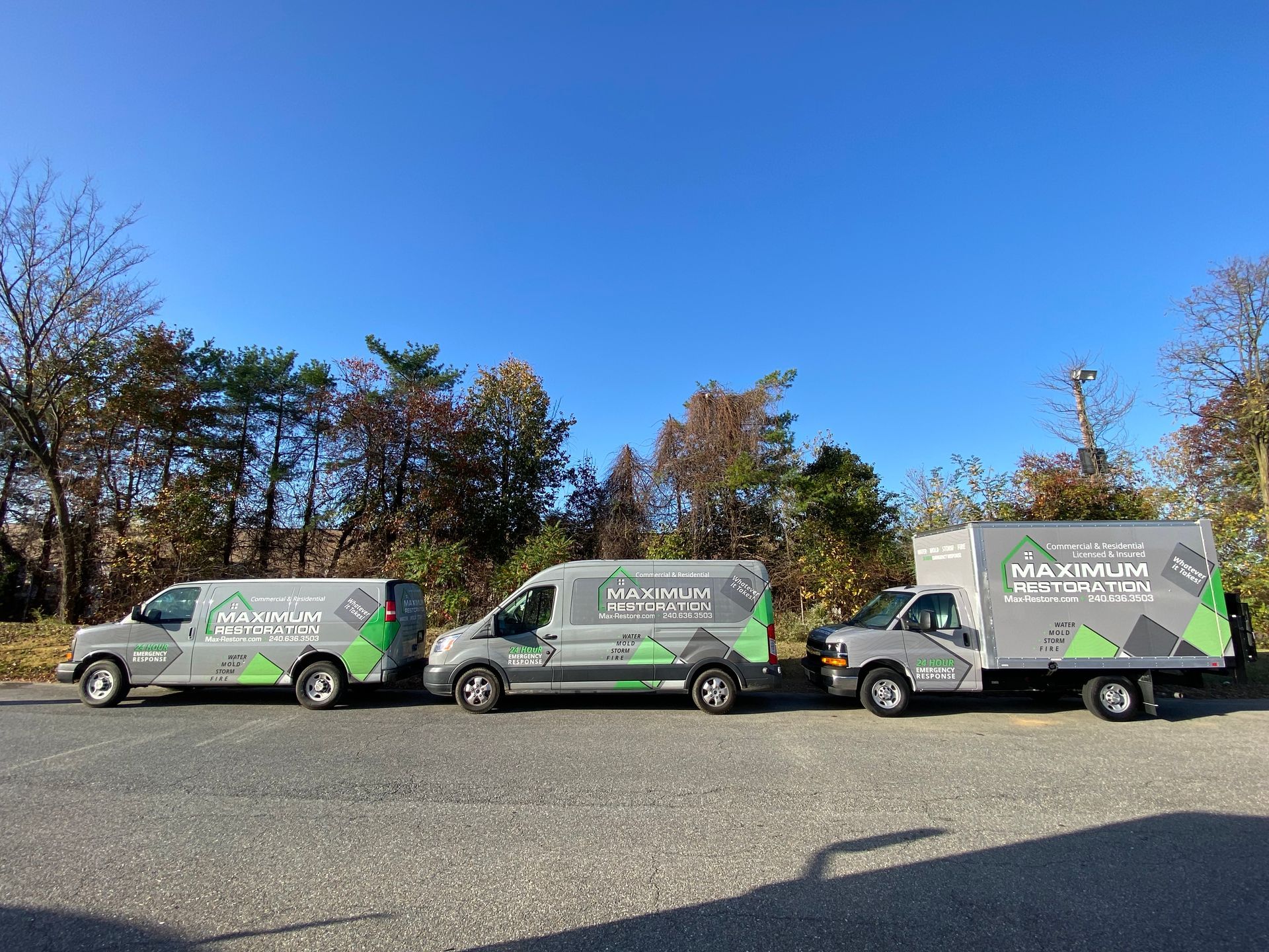 Three gray work vans with green logos parked under a blue sky, trees in the background.