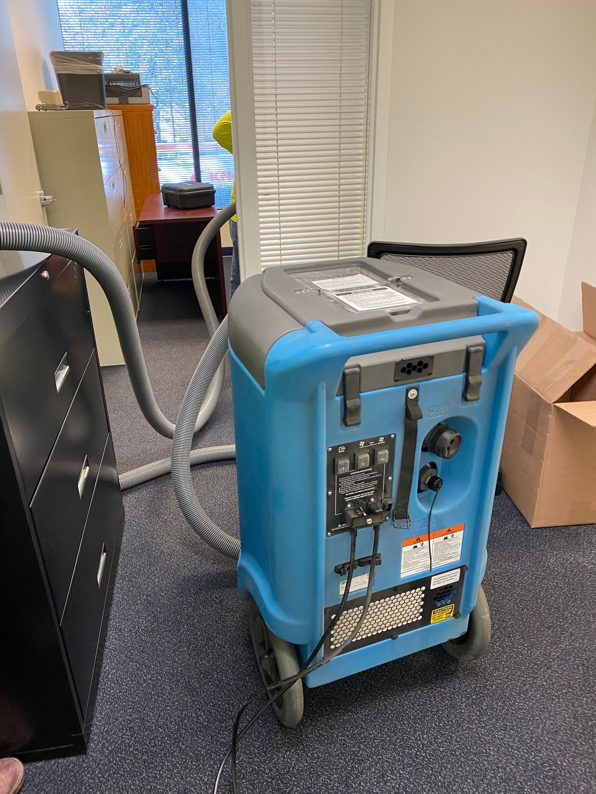 Blue and gray industrial machine with controls, connected to a black cabinet, indoors with office furniture visible.