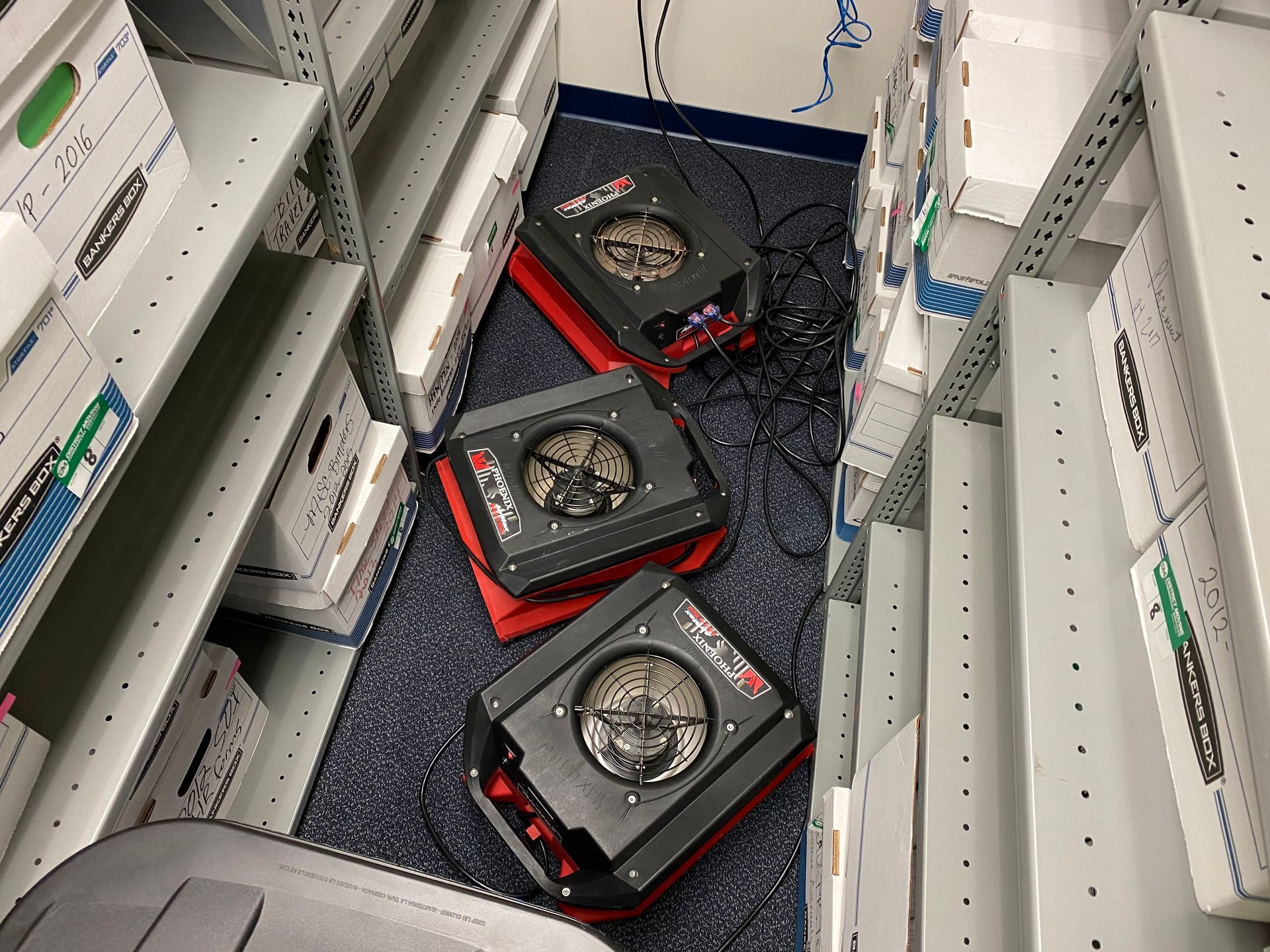 Three square audio speakers on a rug in a storage room with shelves of boxes.