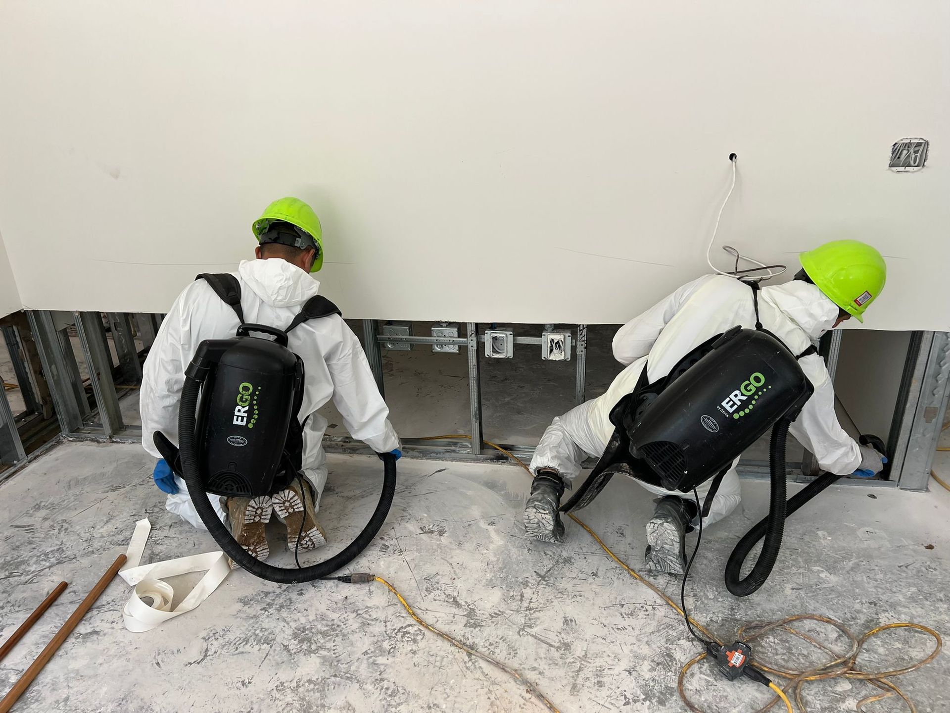 Two workers in protective suits vacuuming debris from a wall in a construction site.