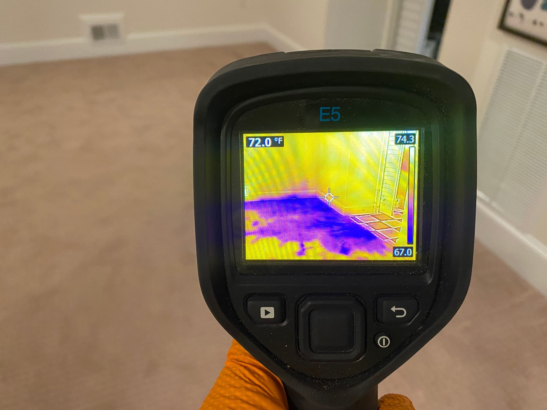 Thermal camera displaying a purple-blue area on a wall, indicating a temperature difference, held by gloved hand.