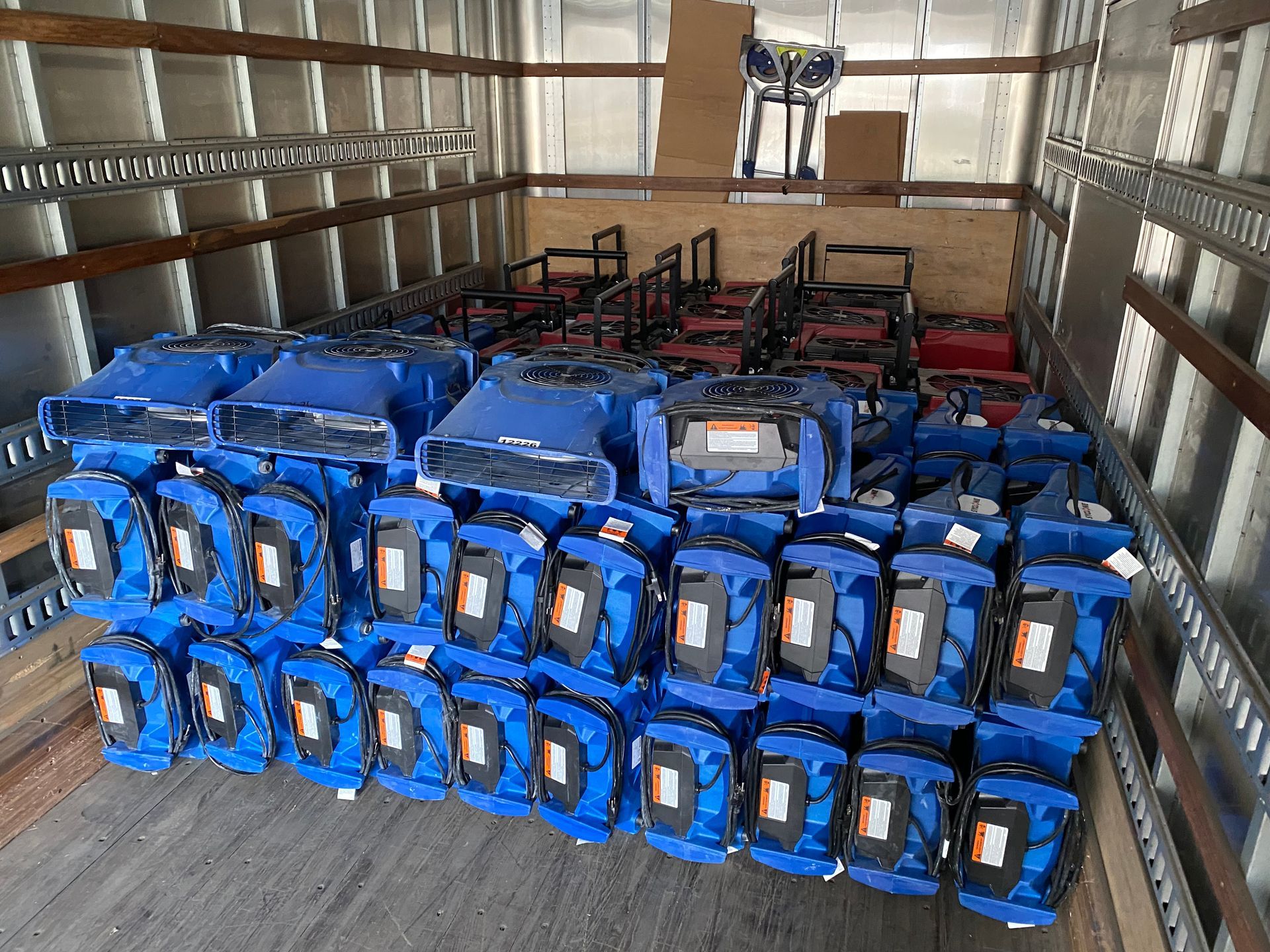 Inside a truck, numerous blue generators and tool bags are stacked against a wall.