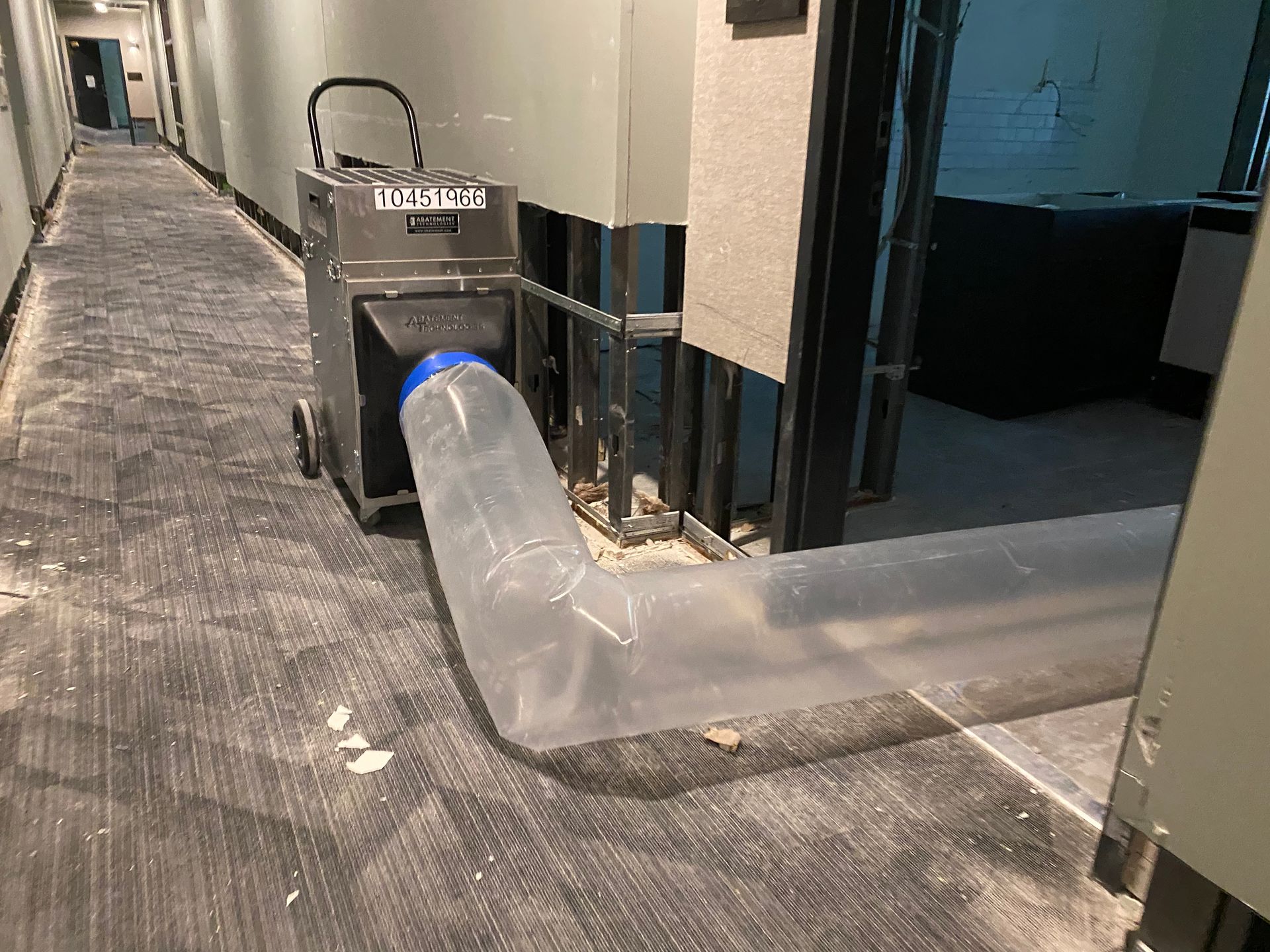 Dehumidifier machine with clear tube in hallway, likely drying water damage. Gray walls, carpet.