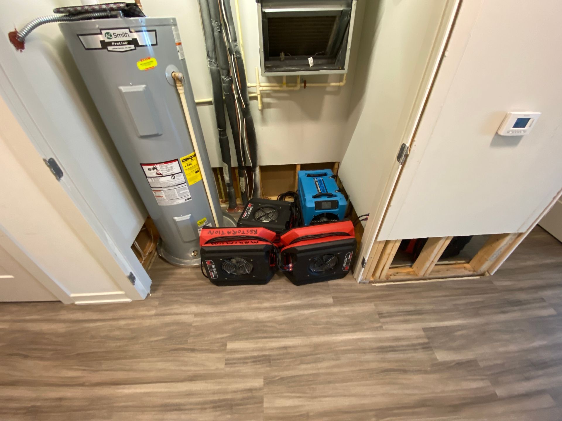 Water heater, HVAC equipment, and tools in a utility closet with exposed wall. Gray flooring.