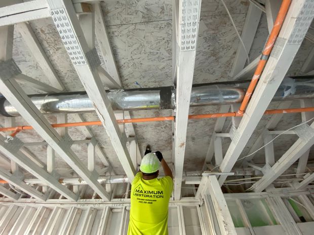 Construction worker drills into a wooden beam, orange pipes and ductwork visible above, inside a building.