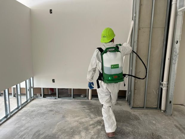 Person in protective suit spraying wall in unfinished room.