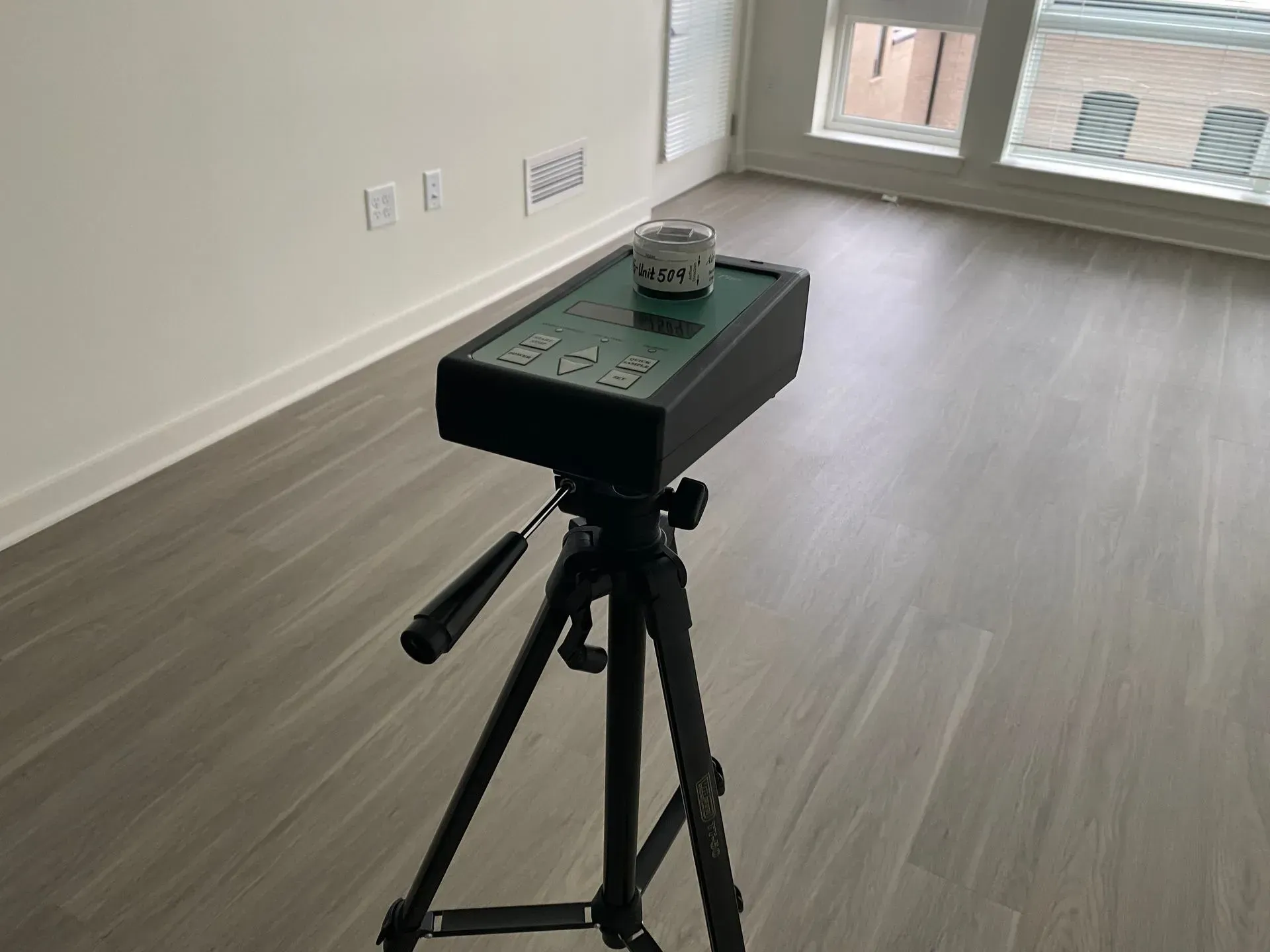 A tripod-mounted environmental monitoring device sits on a light-colored wooden floor in an empty room.