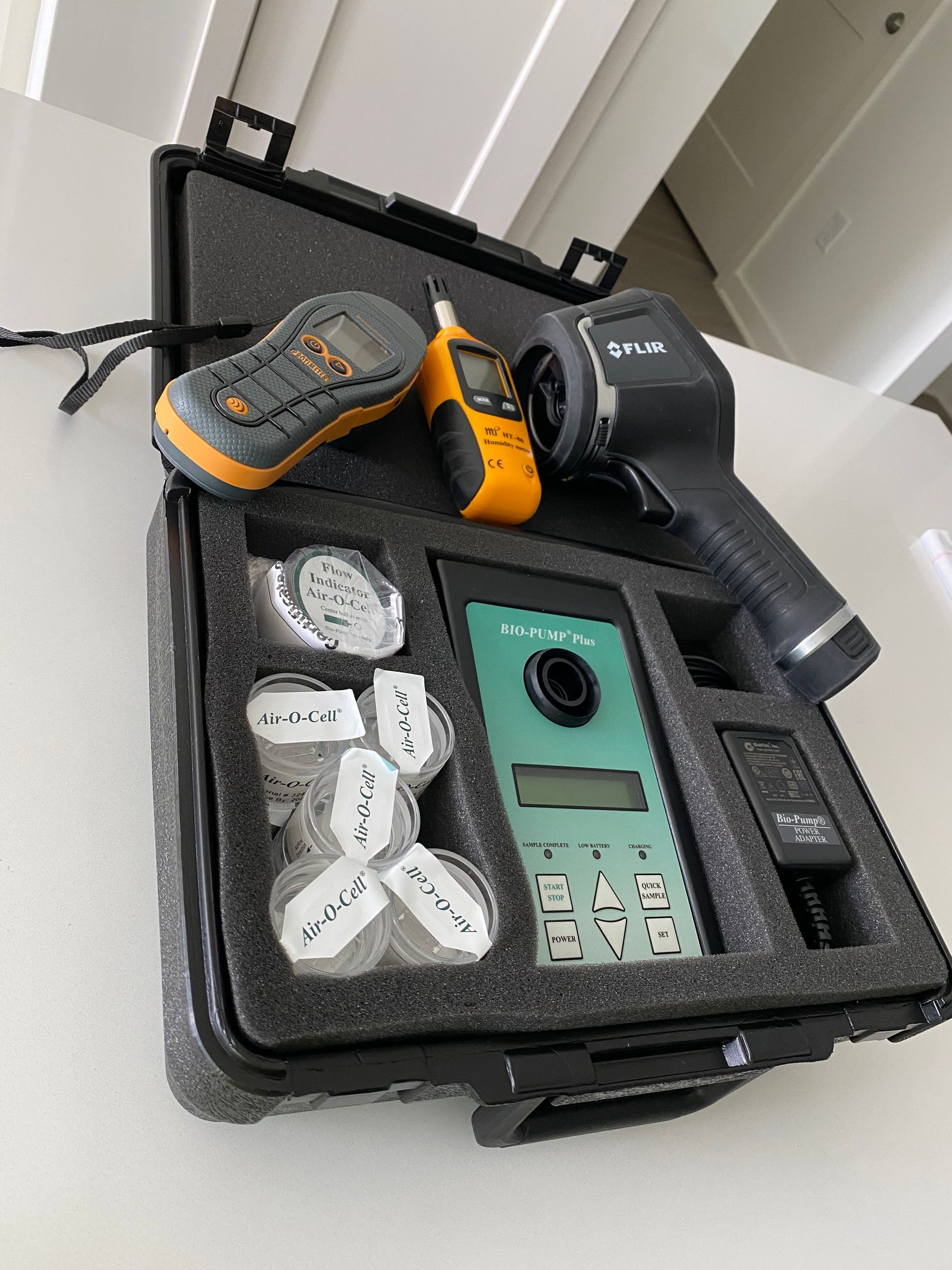 A black hard-shell case containing a FLIR thermal camera, two smaller yellow testing devices, and various sample vials.