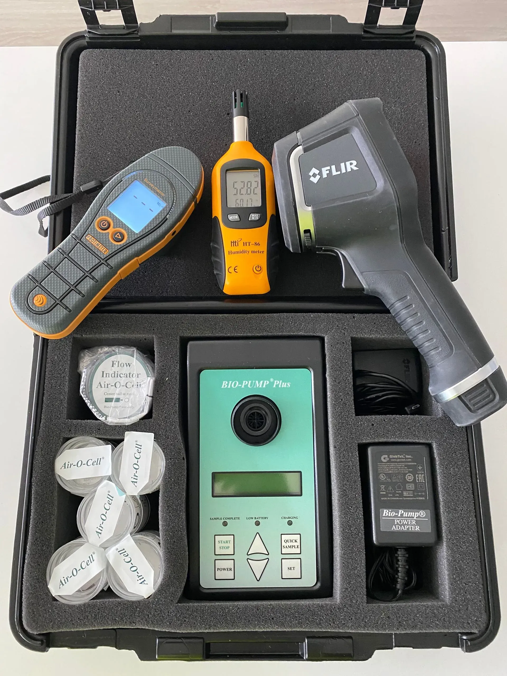 A black carrying case contains a FLIR thermal camera, a handheld meter, a central testing device, and small labeled vials.