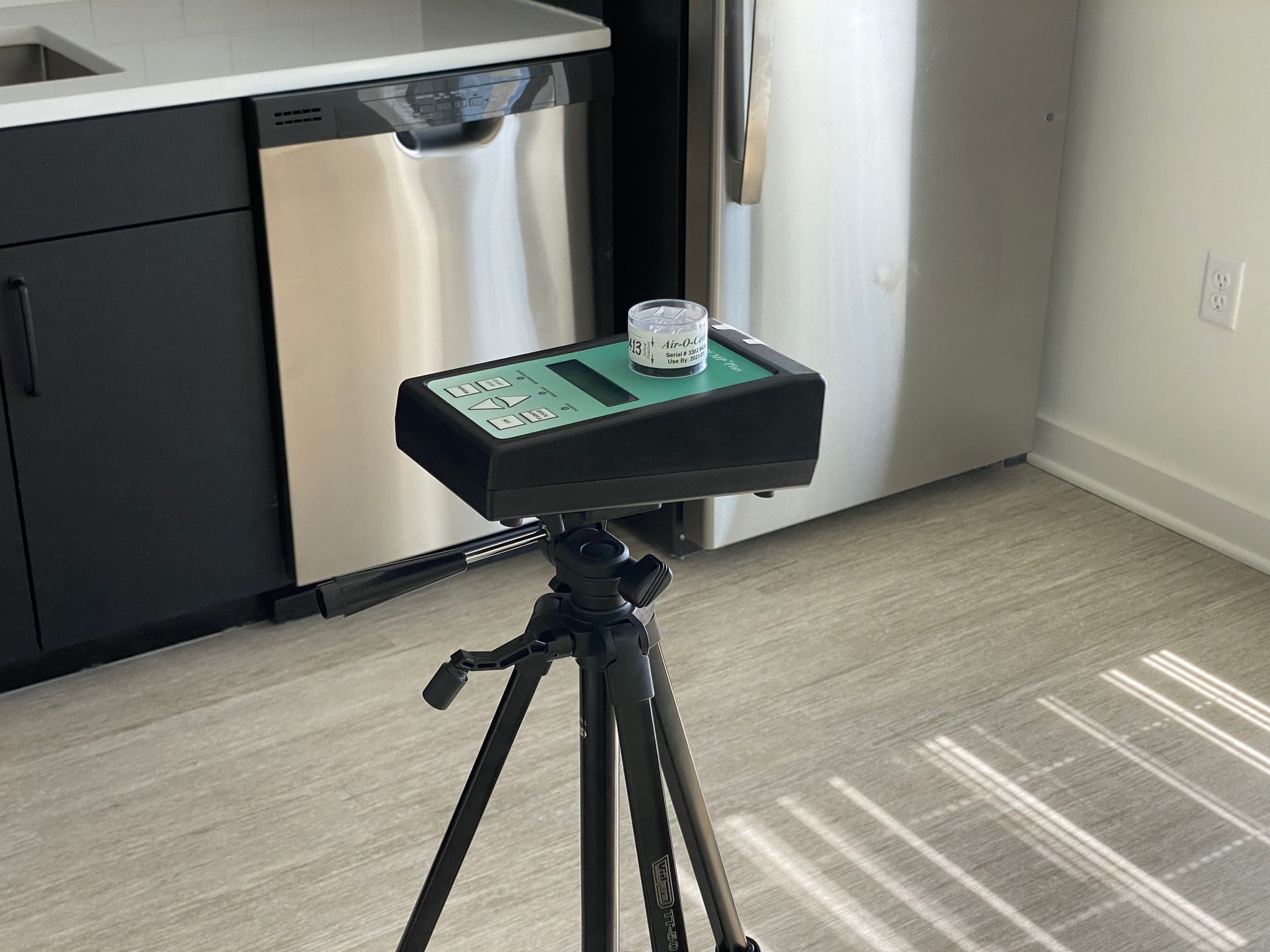 A radon detector on a tripod in a modern kitchen with black cabinets, a stainless steel dishwasher, and a light floor.