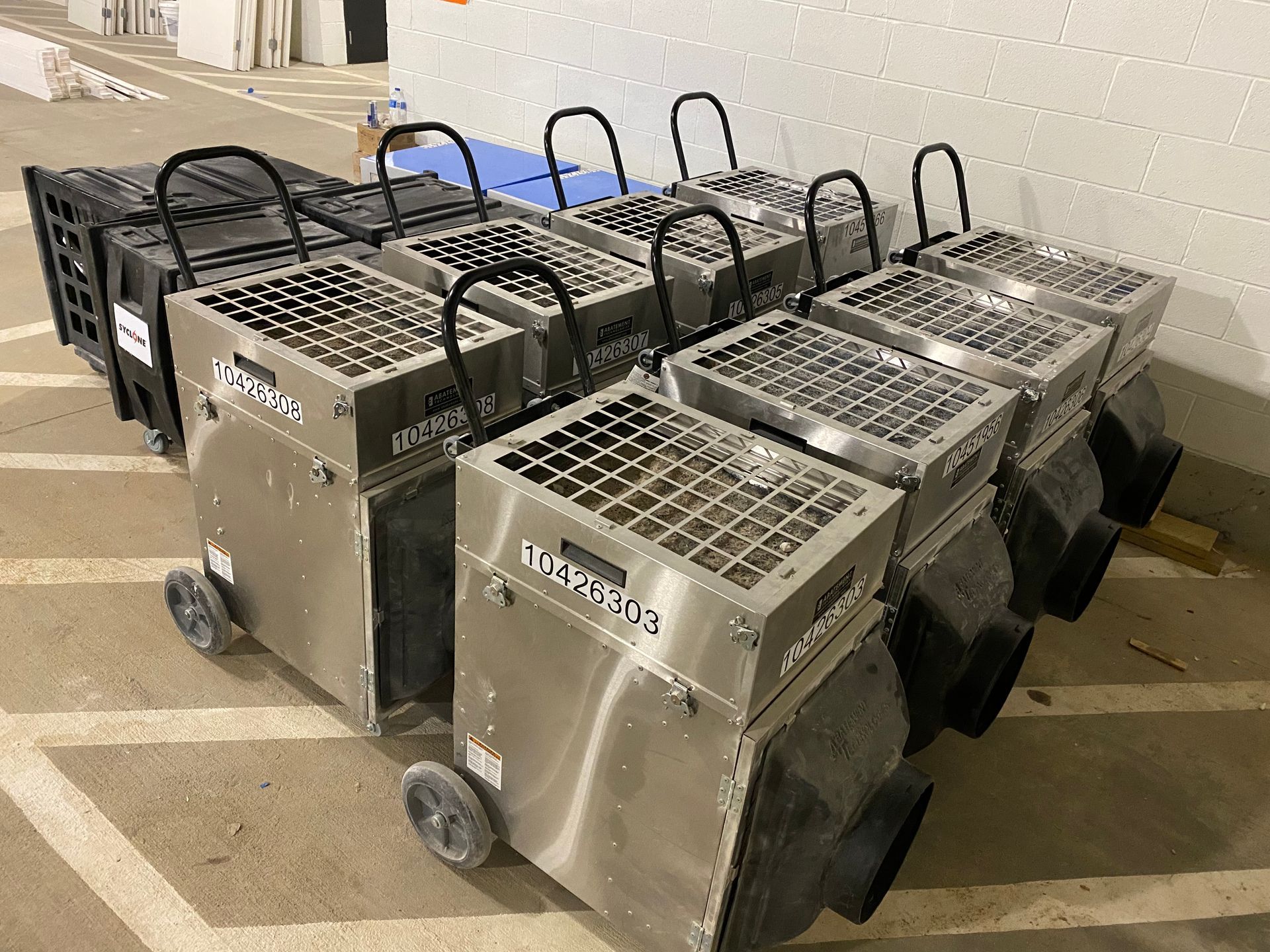 Several metallic, wheeled dehumidifiers lined up in a row on a concrete floor.