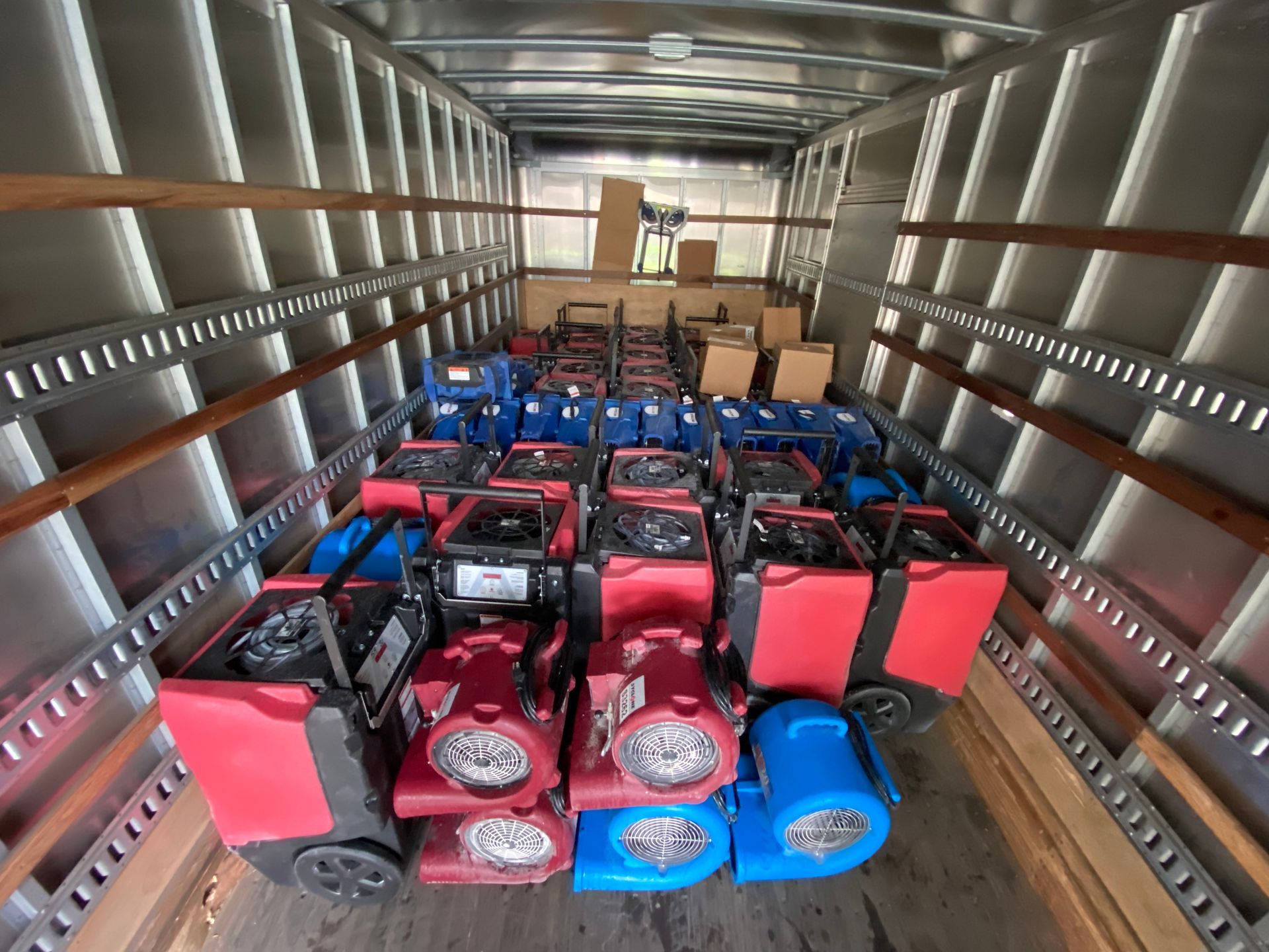 A cargo truck interior filled with rows of stacked industrial air movers and dehumidifiers in red and blue.