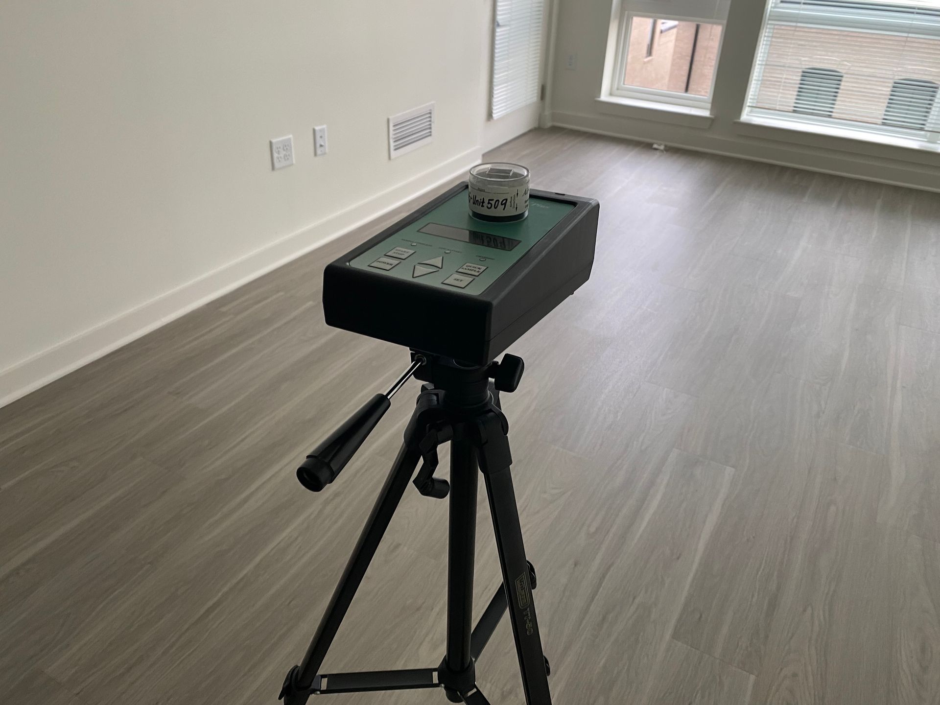 A professional air quality monitor on a tripod stands in the center of an empty, light-filled room with wood-style floors.
