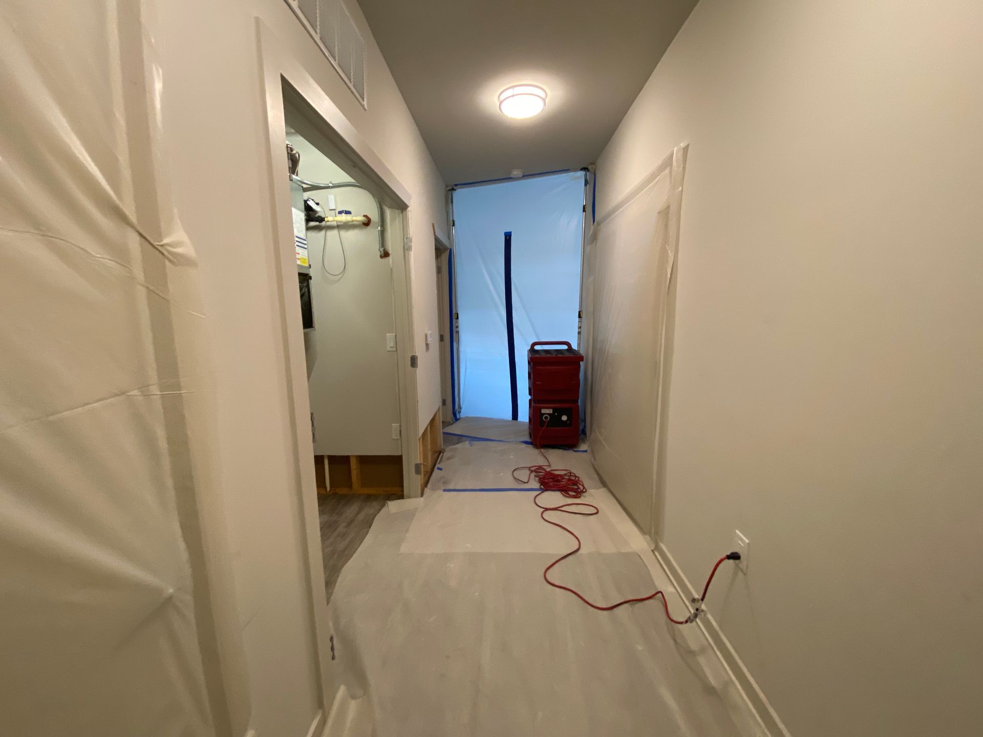 A hallway under construction with protective plastic sheeting on the walls, floor coverings, and a red dehumidifier.