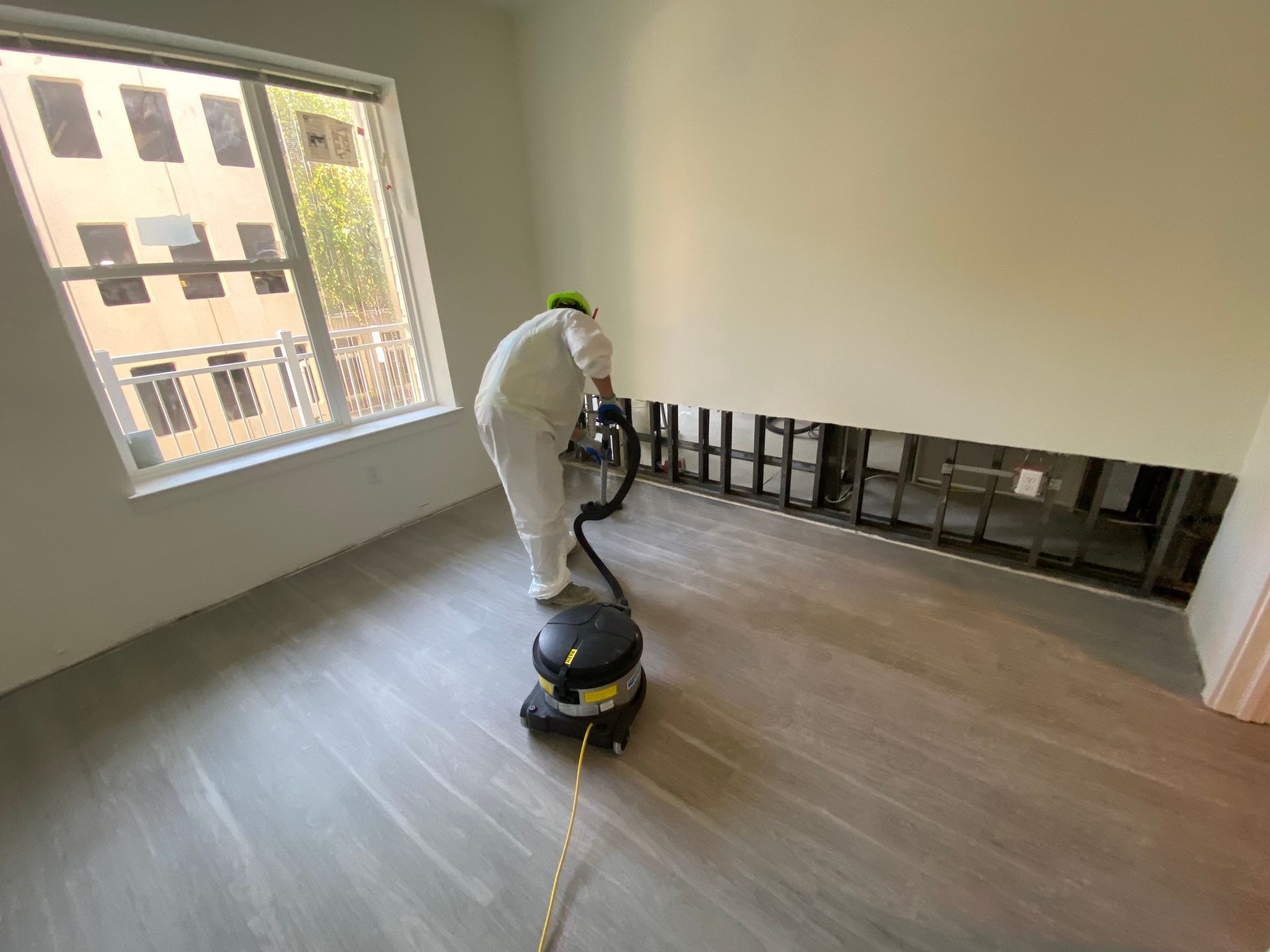 A person in protective coveralls vacuums a room where a section of the lower wall has been removed to expose the studs.