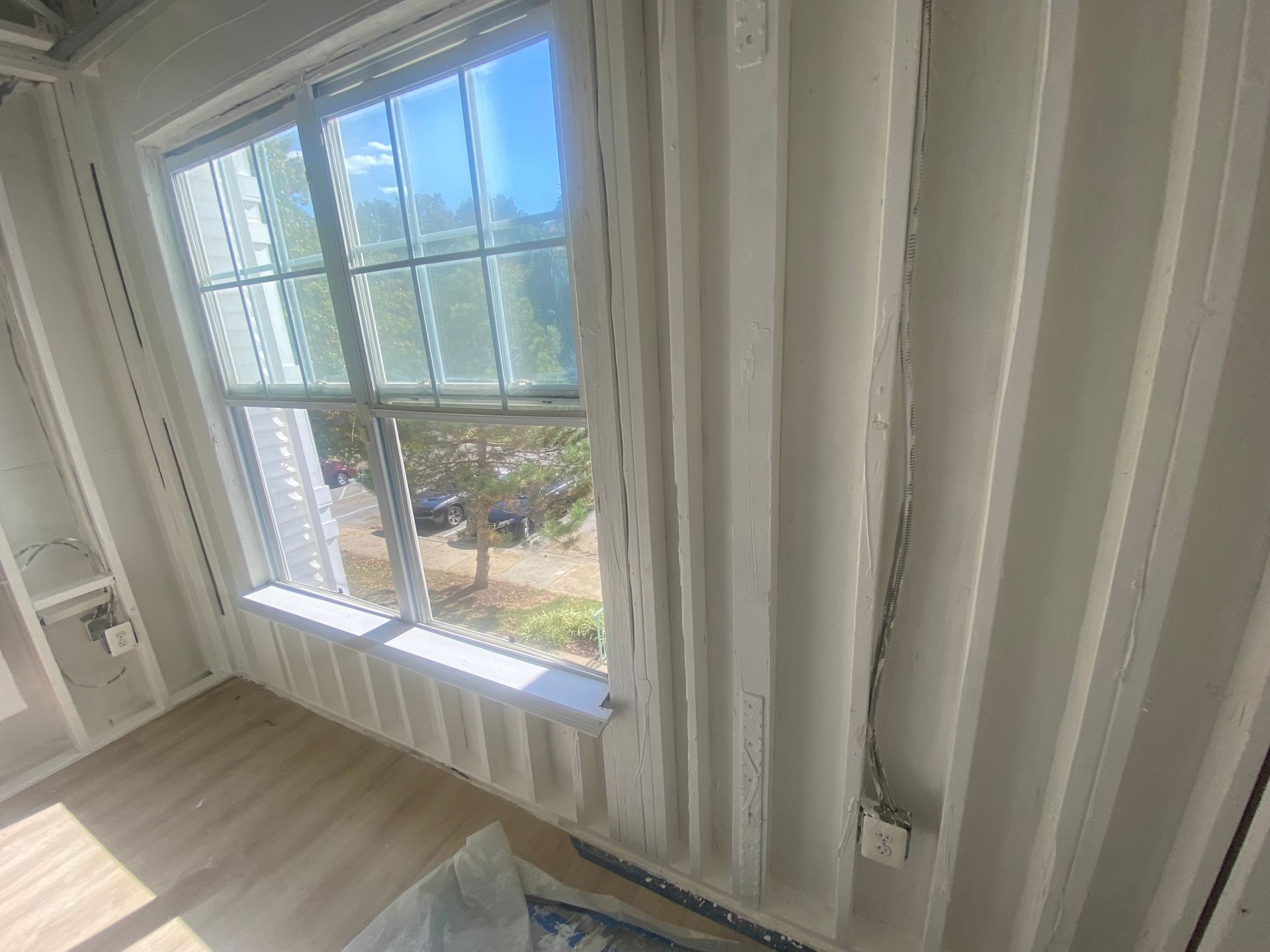 An unfinished room with white wood framing, exposed electrical wiring on a wall, and a large window overlooking trees.