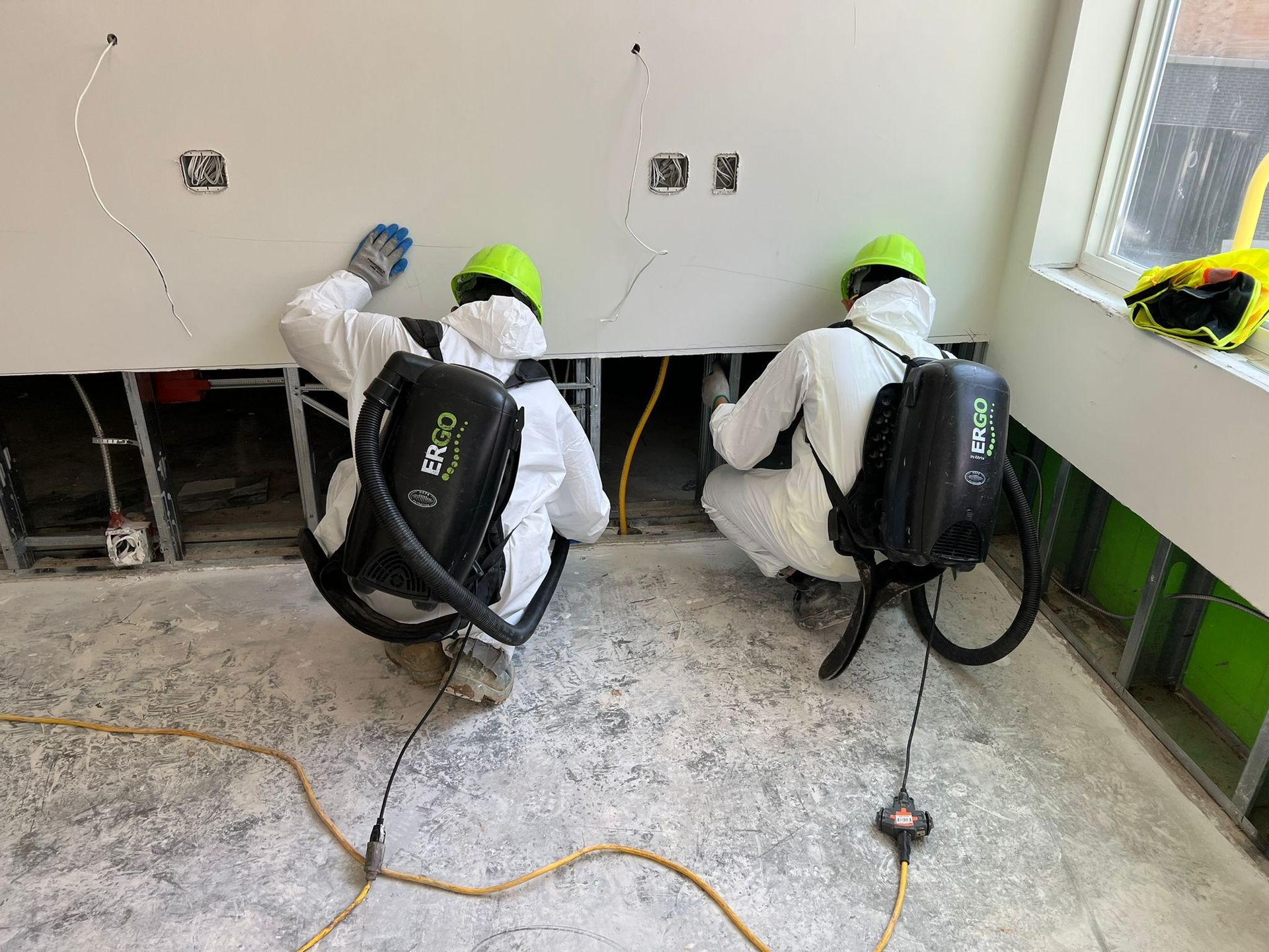 Two workers in white hazmat suits and lime green hard hats use backpack vacuums while kneeling to clean a dusty floor.