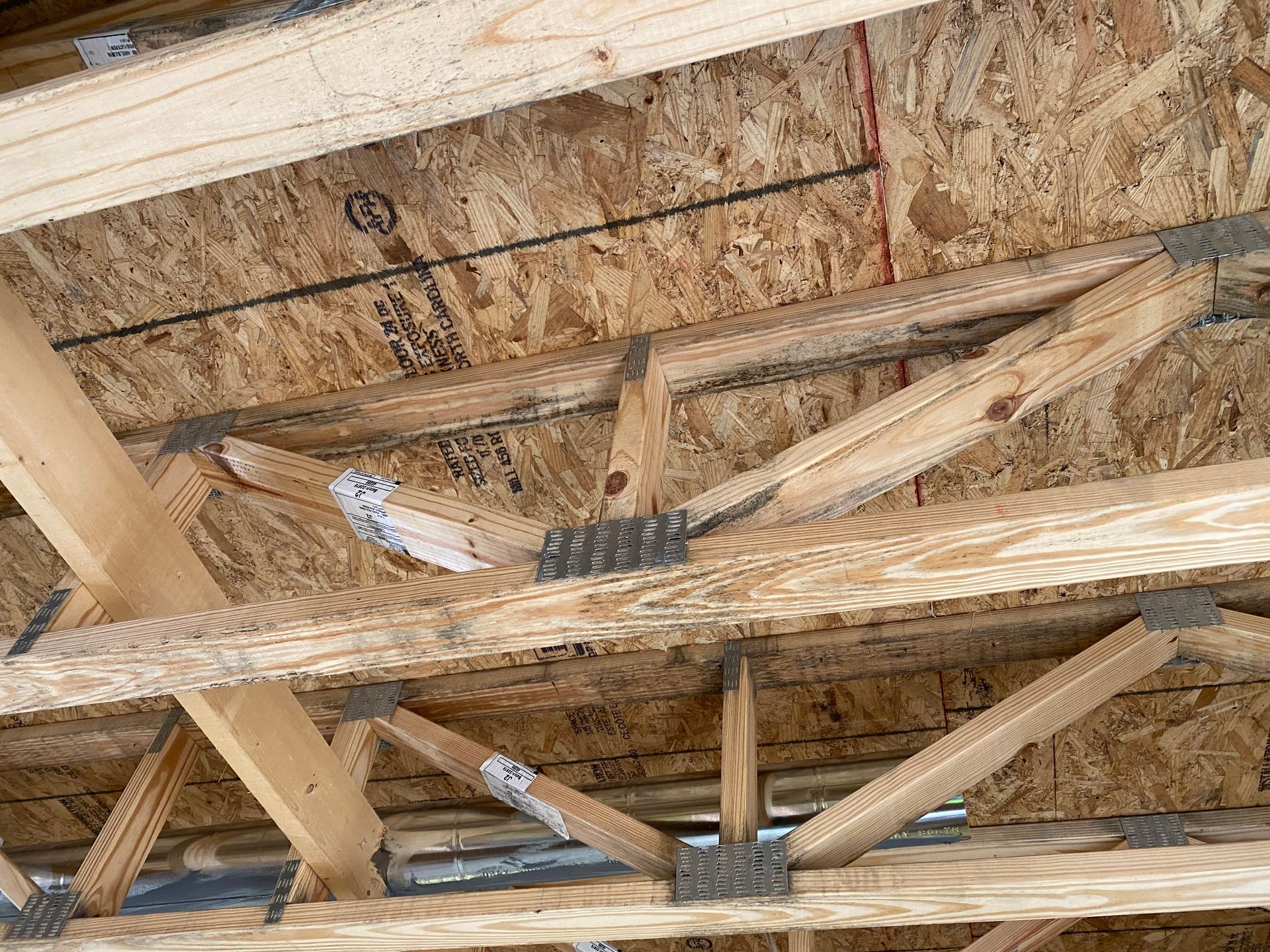 The underside of a wooden roof structure, showing trusses, connecting metal plates, and oriented strand board sheathing.