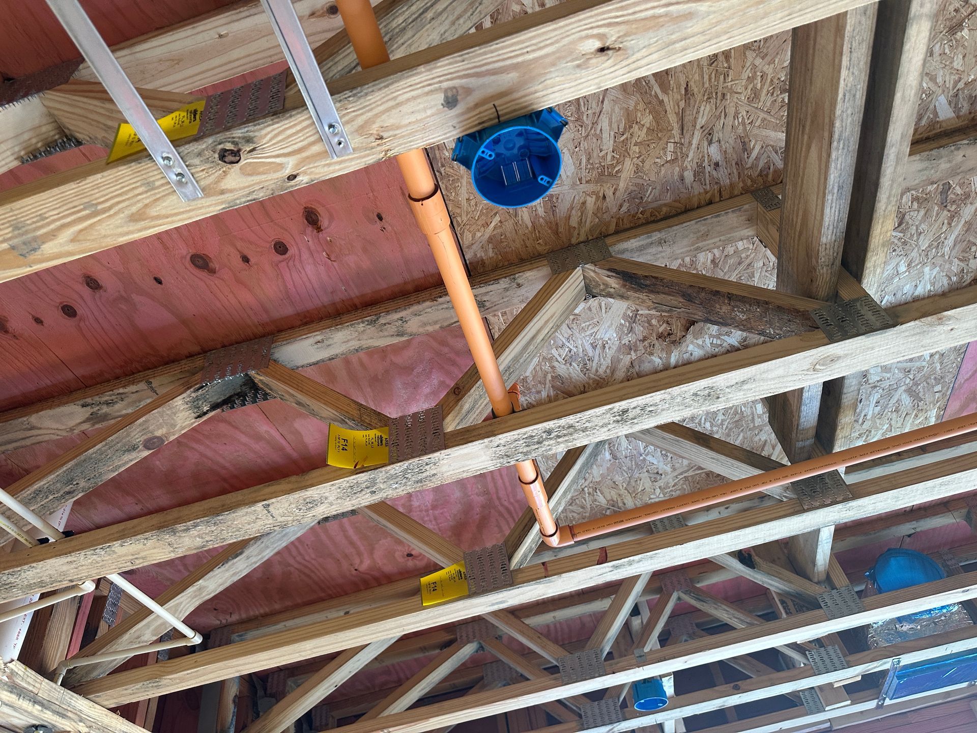 Blue electrical junction boxes and orange conduits installed on wooden ceiling joists in an unfinished room.