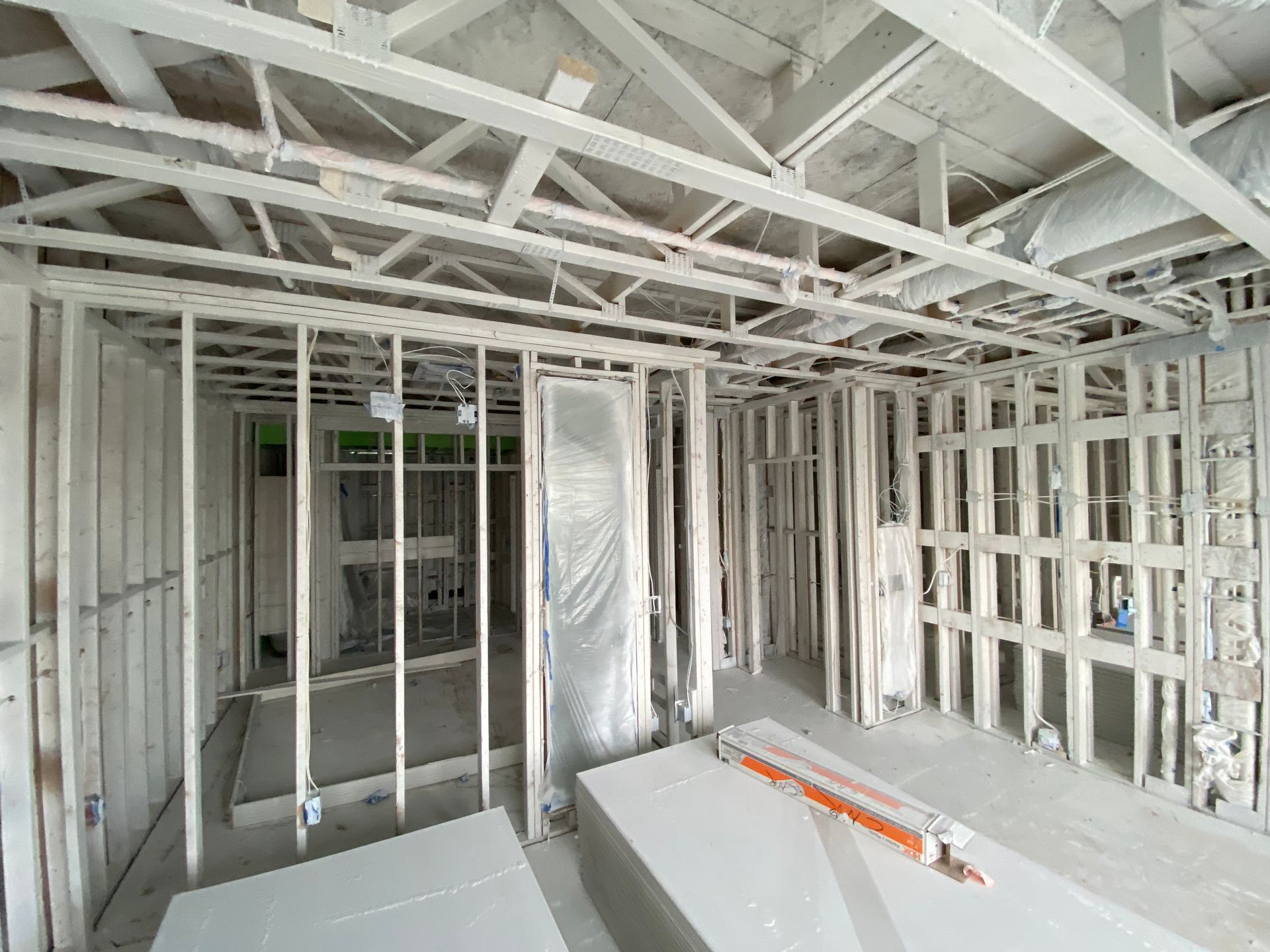 An interior view of a room under construction, showing wooden wall framing, open ceilings, and stacks of drywall.