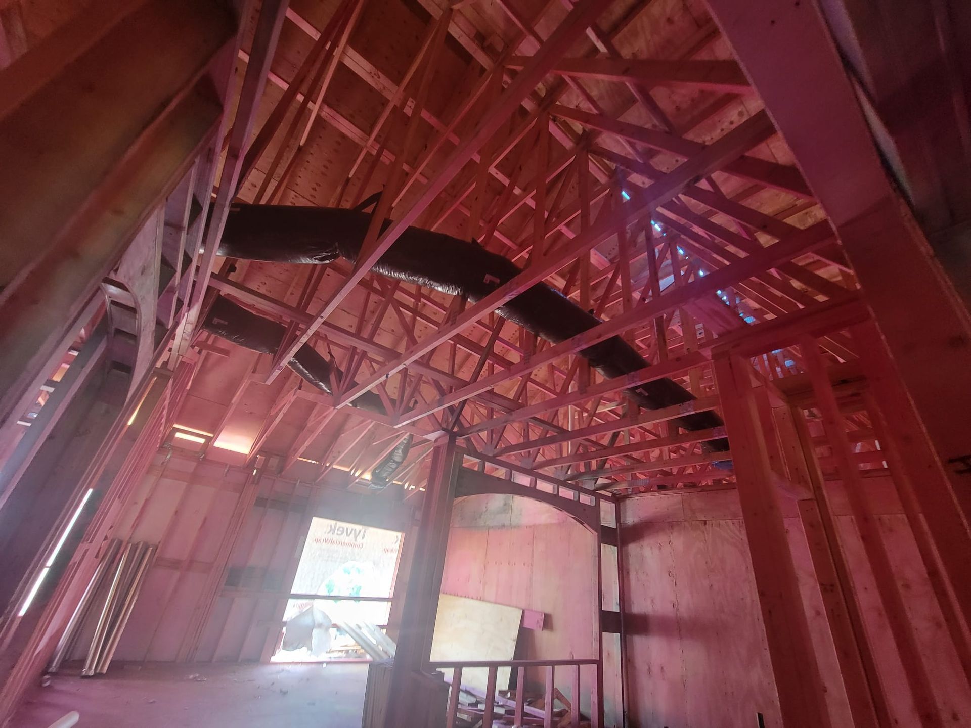 A construction site interior showing wooden framing, roof trusses, and red-tinted spray foam insulation on the walls.