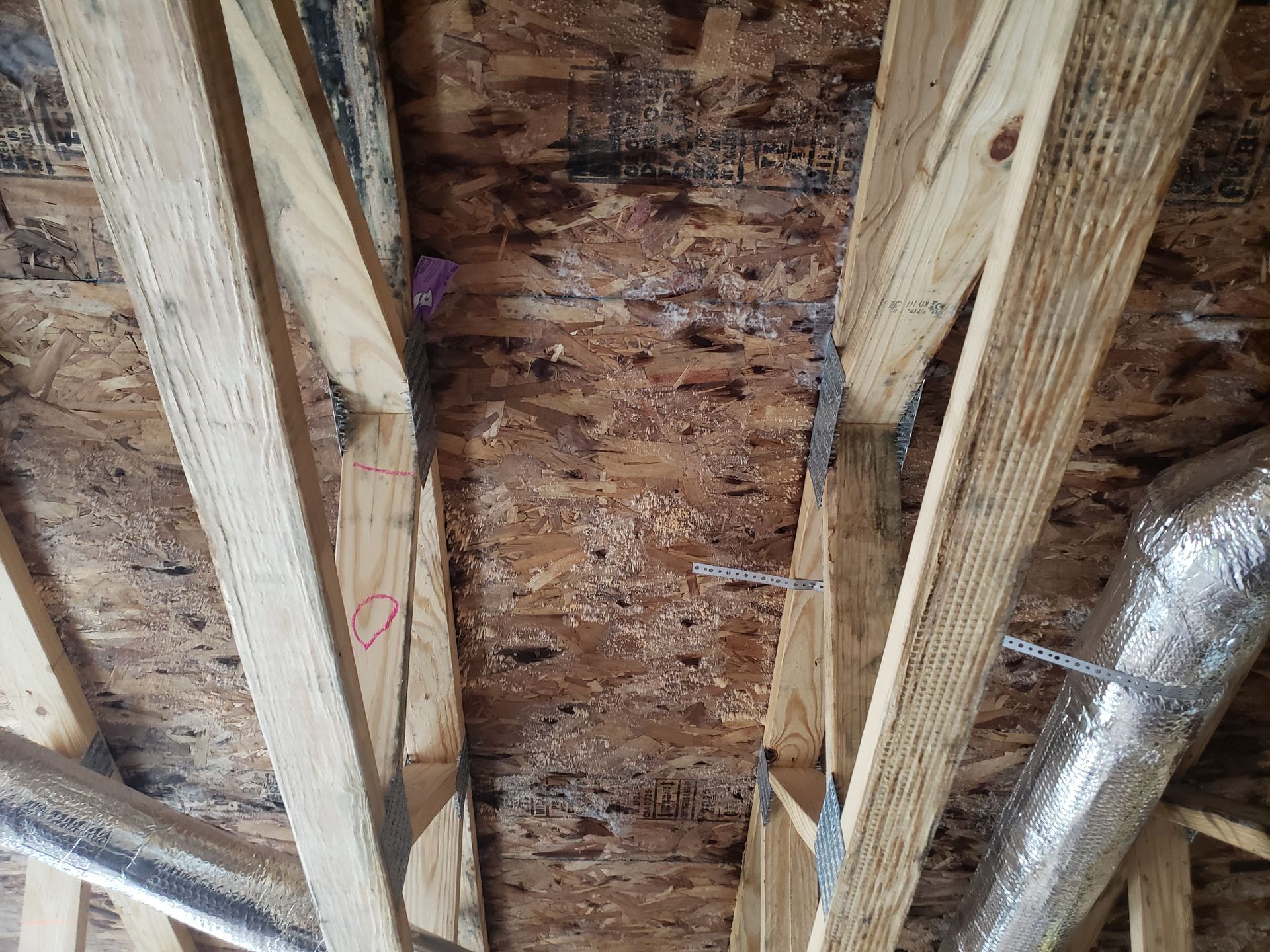View from inside an attic showing wooden roof trusses, plywood sheathing with dark stains, and metal ductwork.