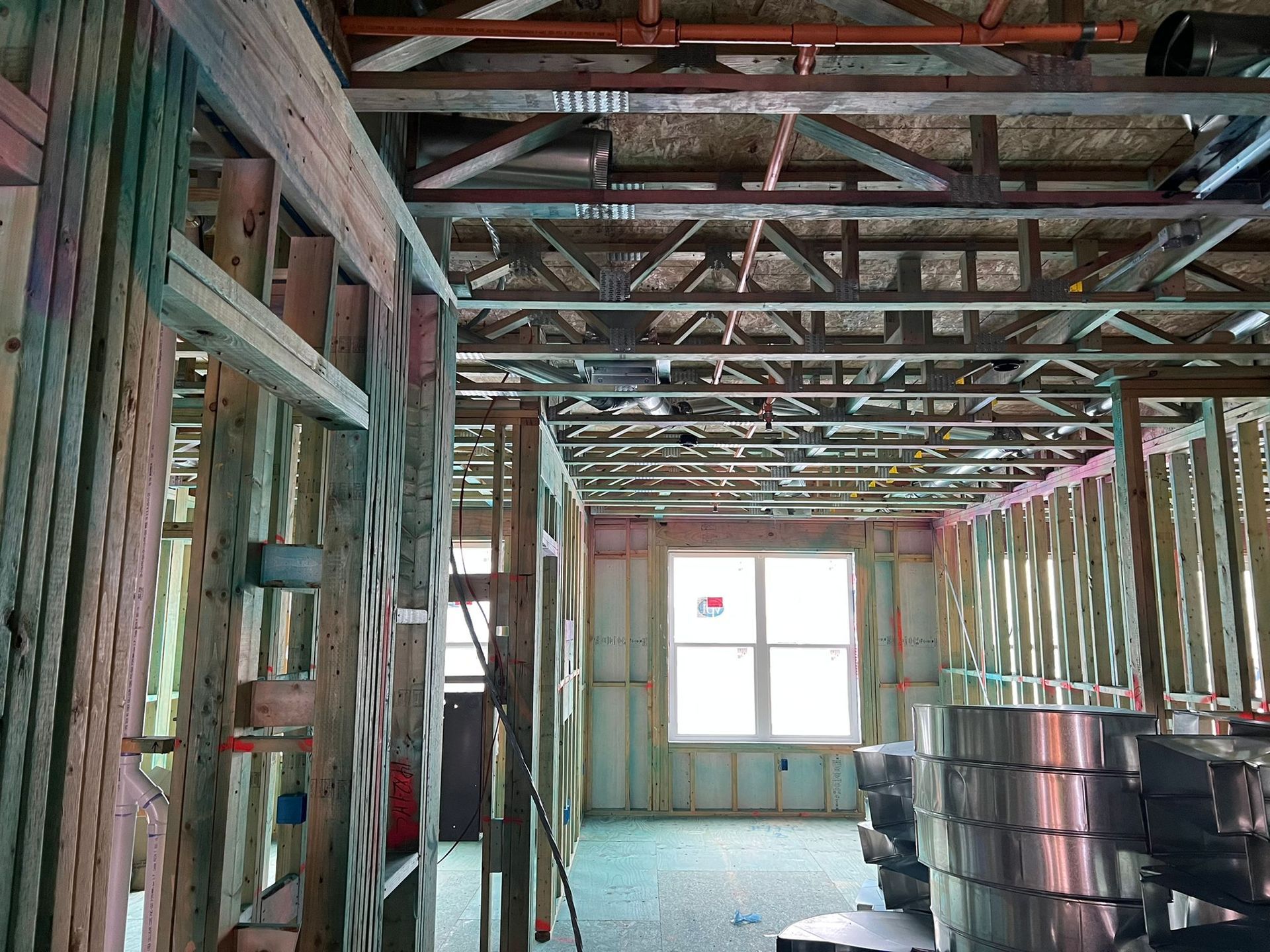 An interior view of a room under construction, showing wooden wall framing, open metal roof trusses, and a window.