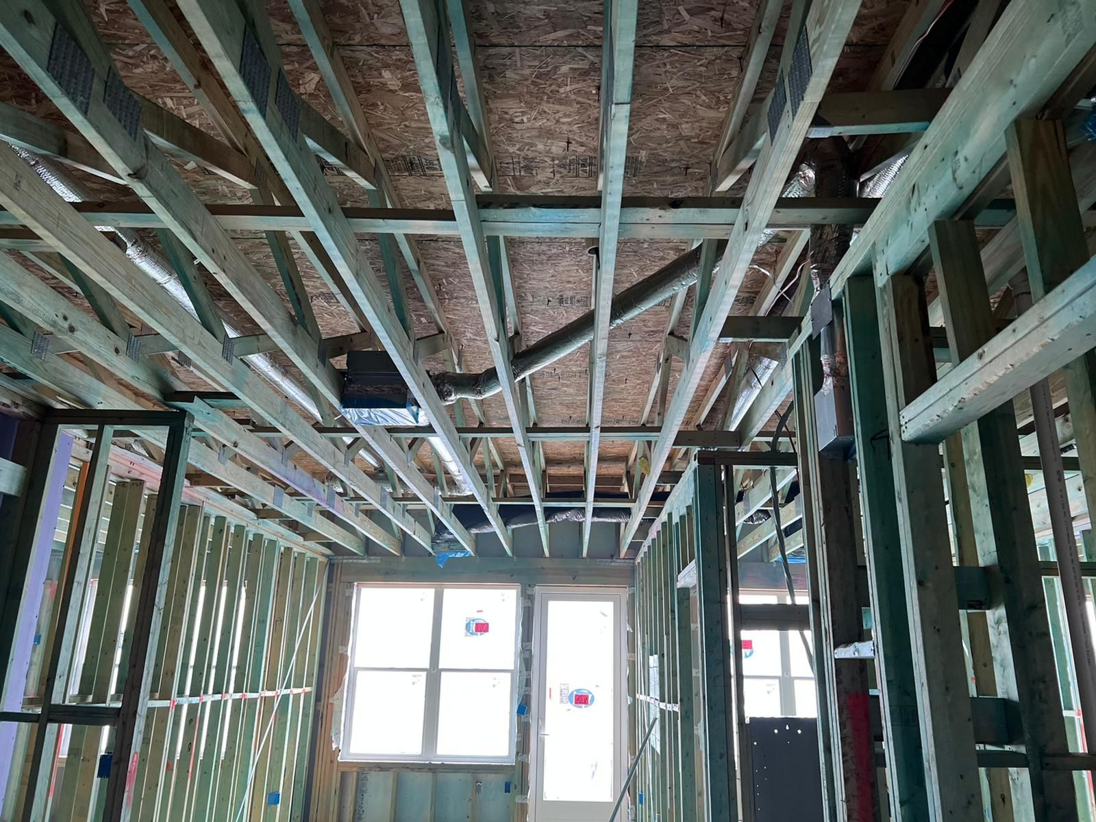 Exposed wooden framing and ceiling joists inside a house under construction, with windows at the far wall.