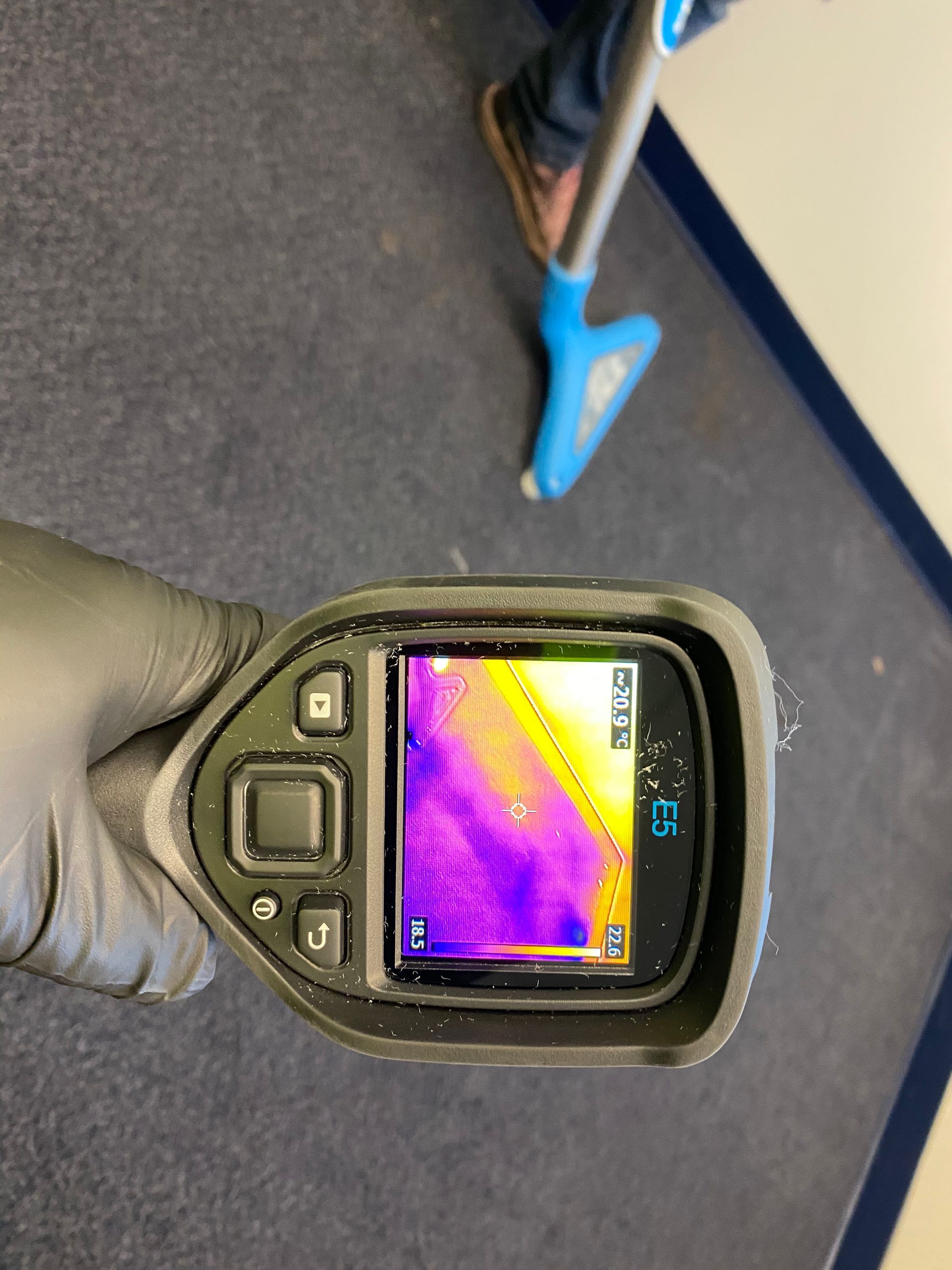 A gloved hand holds a thermal imaging camera showing a heat gradient on a carpeted floor near a cleaning tool.