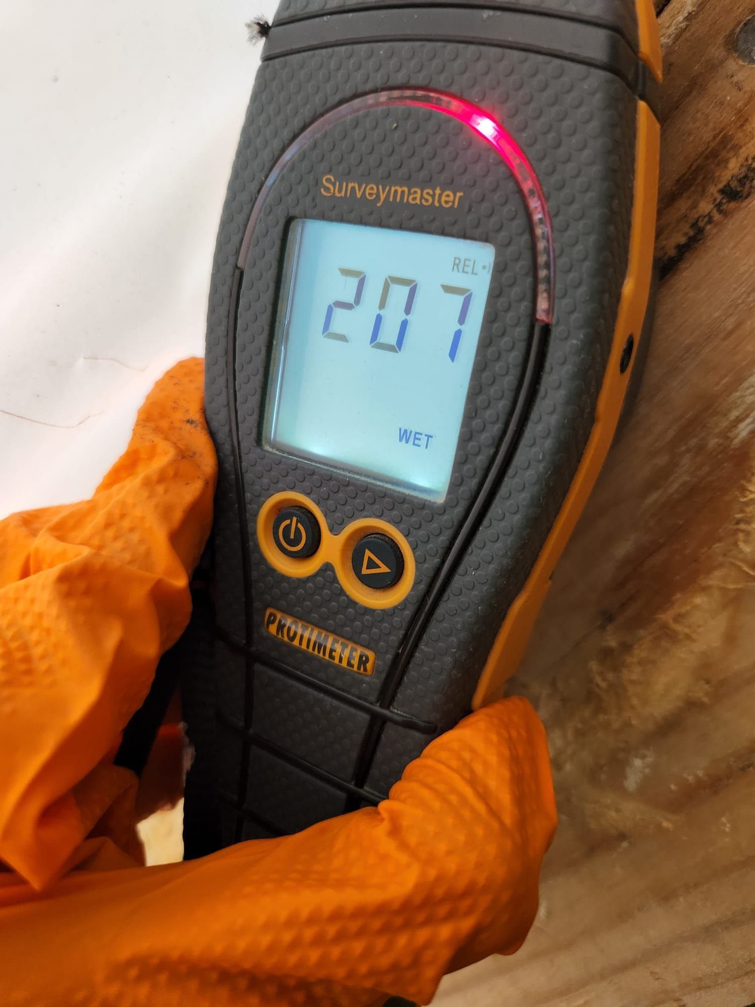 Gloved hand holding a digital moisture meter against a wooden surface, displaying a reading of 207 with a red light.