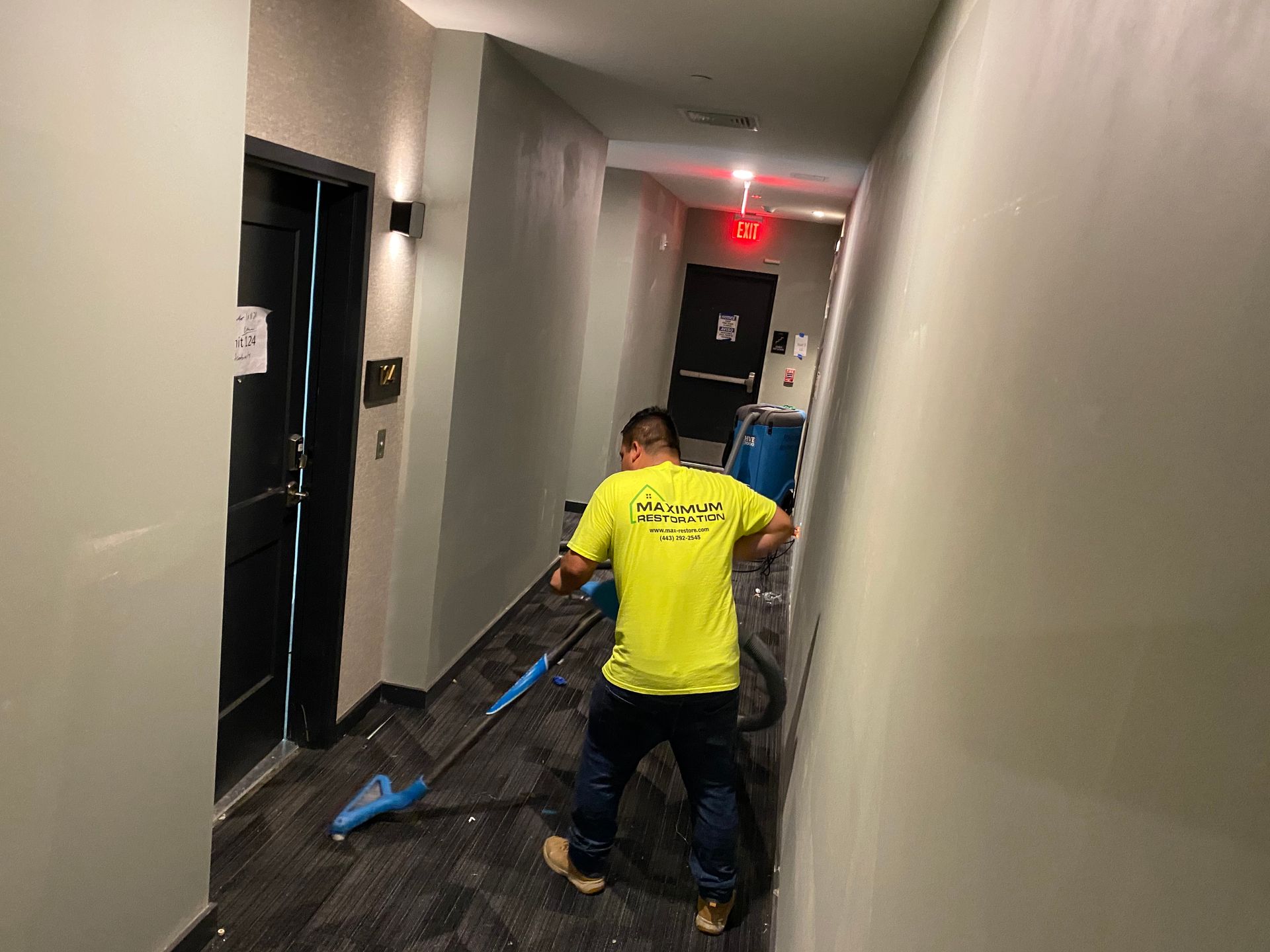 A worker in a bright yellow shirt uses a carpet cleaning machine in a hotel hallway with dark flooring and light walls.